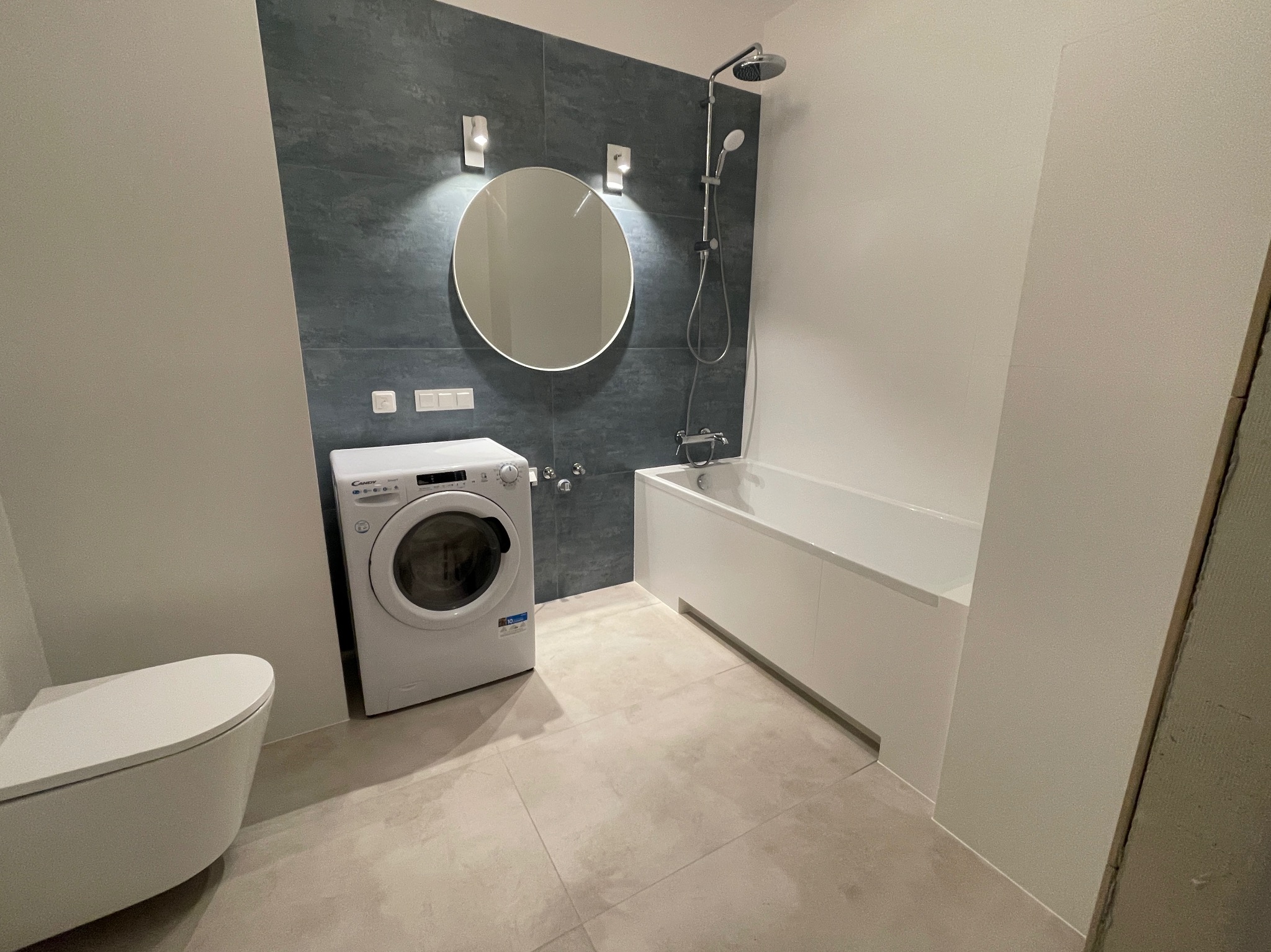 Nowoczesna łazienka z szarą płytką imitującą kamień, okrągłym lustrem podświetlanym od góry, białą wanną, toaletą i pralką Candy.