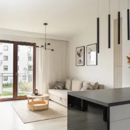 Jasne wnętrze mieszkania w stylu minimalistycznym z beżową sofą, okrągłym stolikiem kawowym, czarnymi lampami wiszącymi i dużym oknem balkonowym z widokiem na budynek.