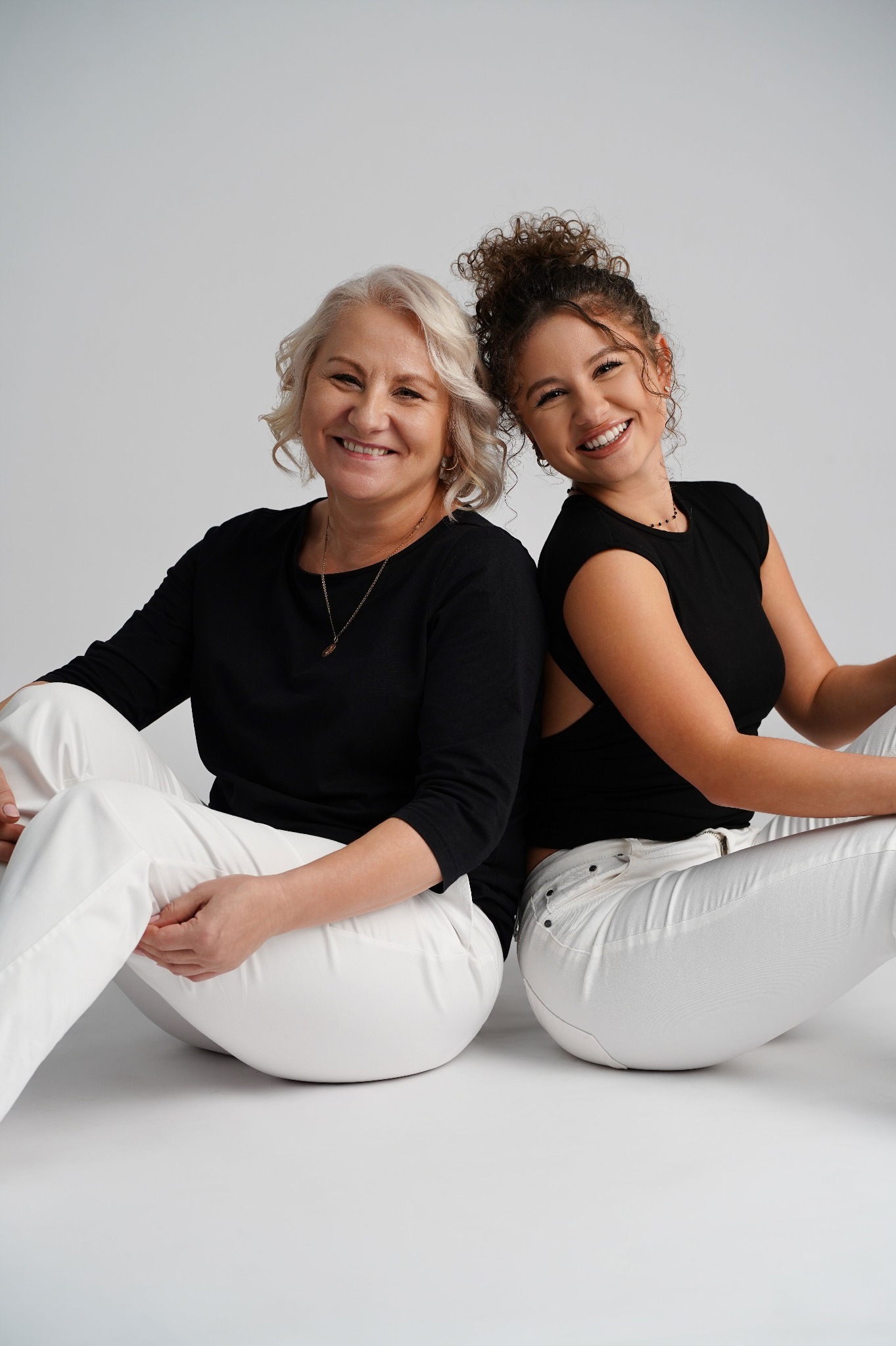 Portret uśmiechniętej matki i córki, siedzących plecami do siebie na białym tle. Obie ubrane w czarne bluzki i białe spodnie, z widocznymi detalami biżuterii.