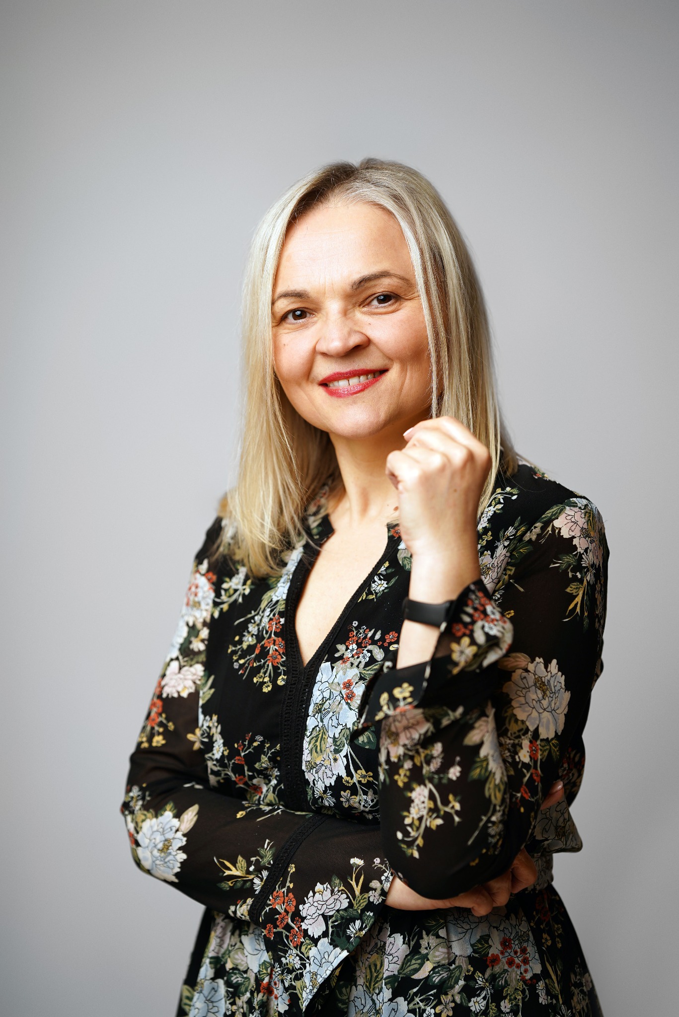 Portret uśmiechniętej kobiety z blond włosami w kwiecistej sukience na szarym tle, z zegarkiem na nadgarstku i rękami skrzyżowanymi na wysokości klatki piersiowej.