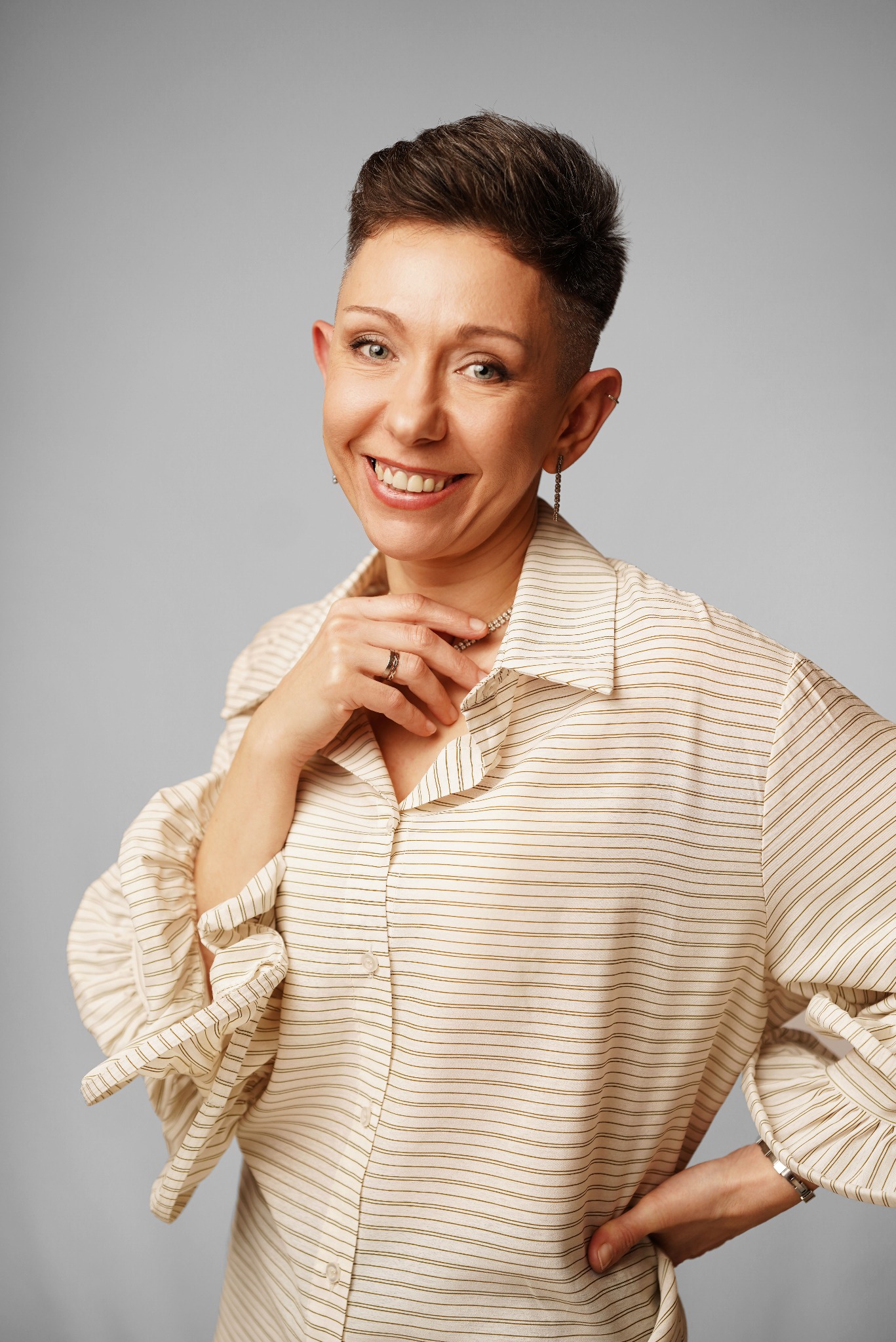 Uśmiechnięta kobieta z krótkimi włosami w koszuli w paski, z ręką na dekolcie i drugą na biodrze, pozuje na jasnoszarym tle. Widoczne kolczyki i pierścionek.
