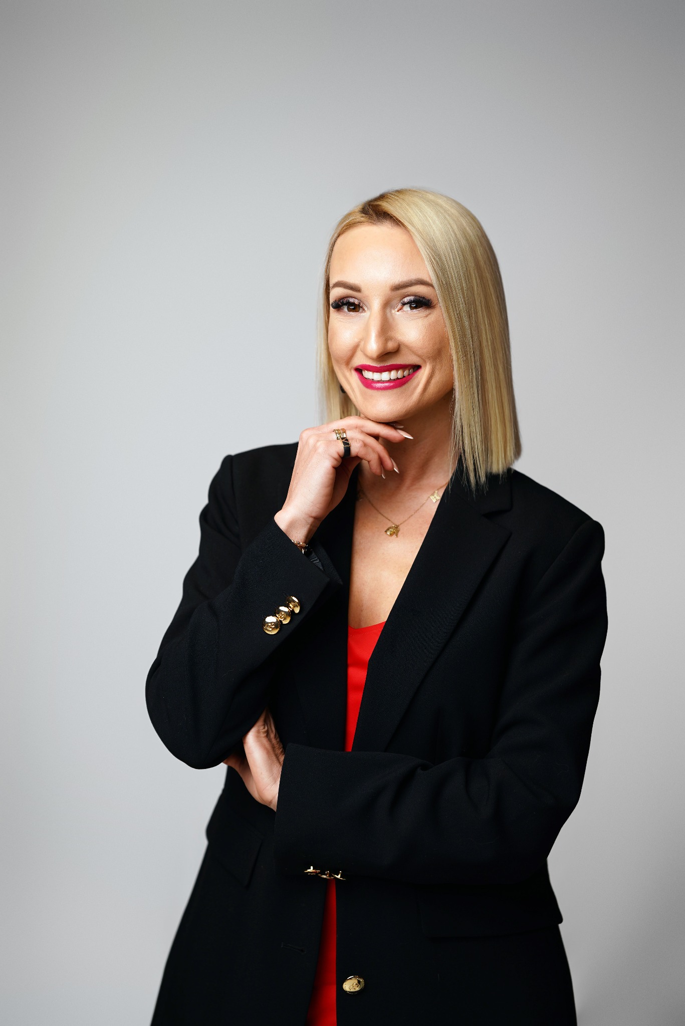 Portret uśmiechniętej blondynki w czarnej marynarce i czerwonej sukience na jasnym tle. Dłoń przy twarzy, biżuteria, profesjonalny makijaż.