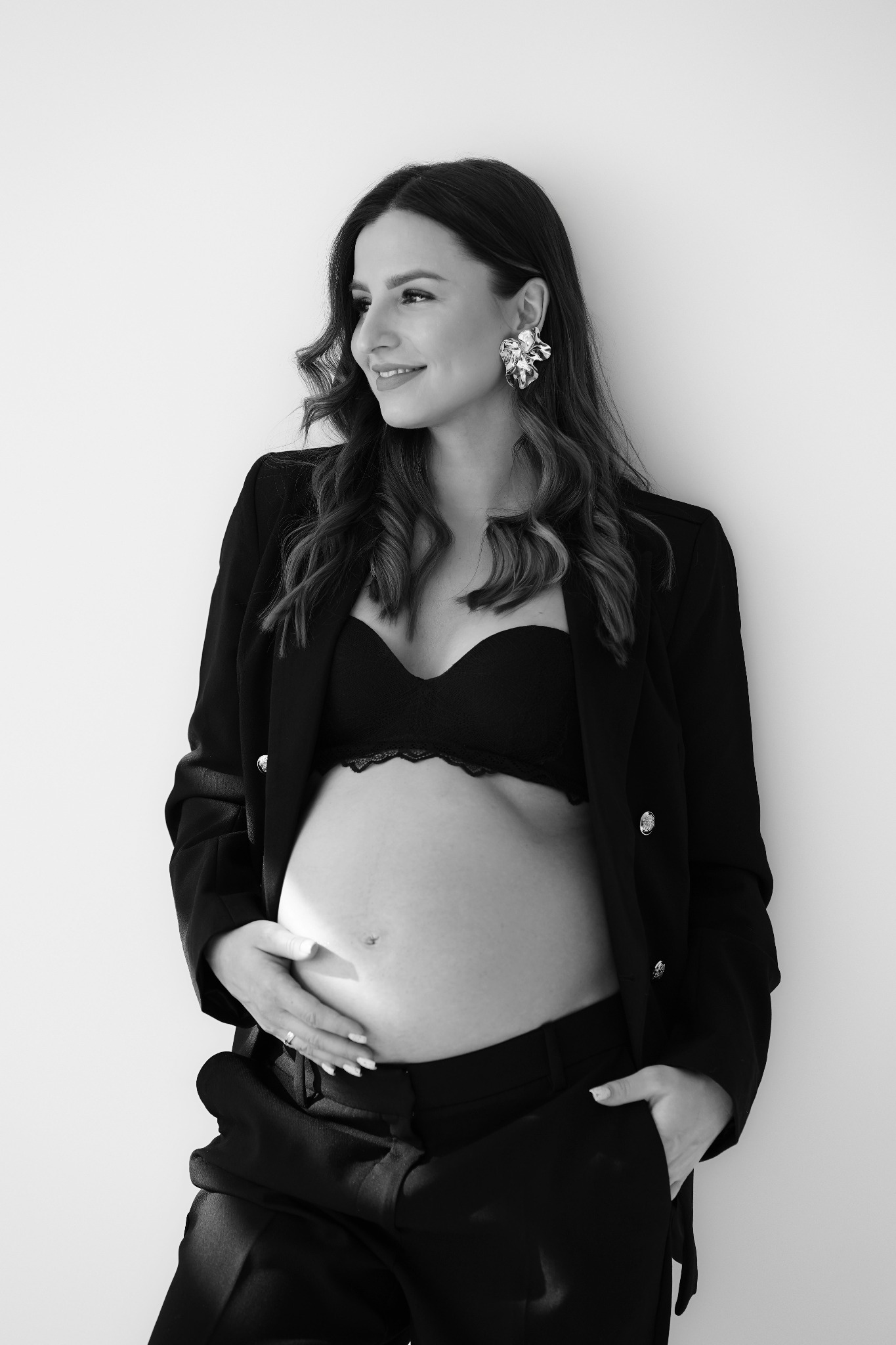 Czarno-białe zdjęcie kobiety w ciąży, ubrana w marynarkę i biustonosz, z ręką na brzuchu, pozująca na jasnym tle. Widoczne duże kolczyki i delikatny uśmiech.