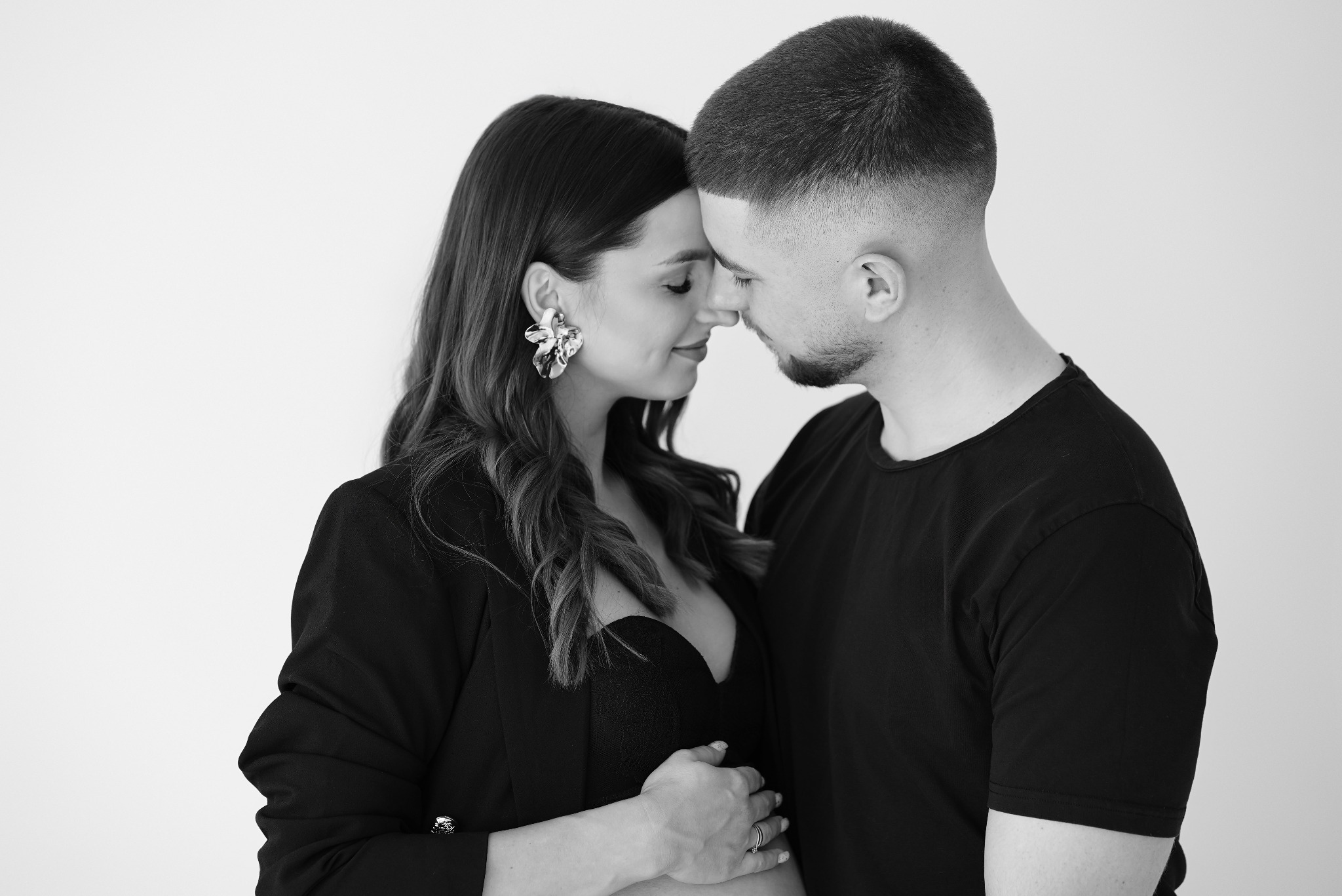 Czarno-białe zbliżenie na parę: kobieta w marynarce i koronkowej bieliźnie, z rękami na brzuchu, dotyka się nosami z mężczyzną w czarnym T-shircie na jasnym tle.