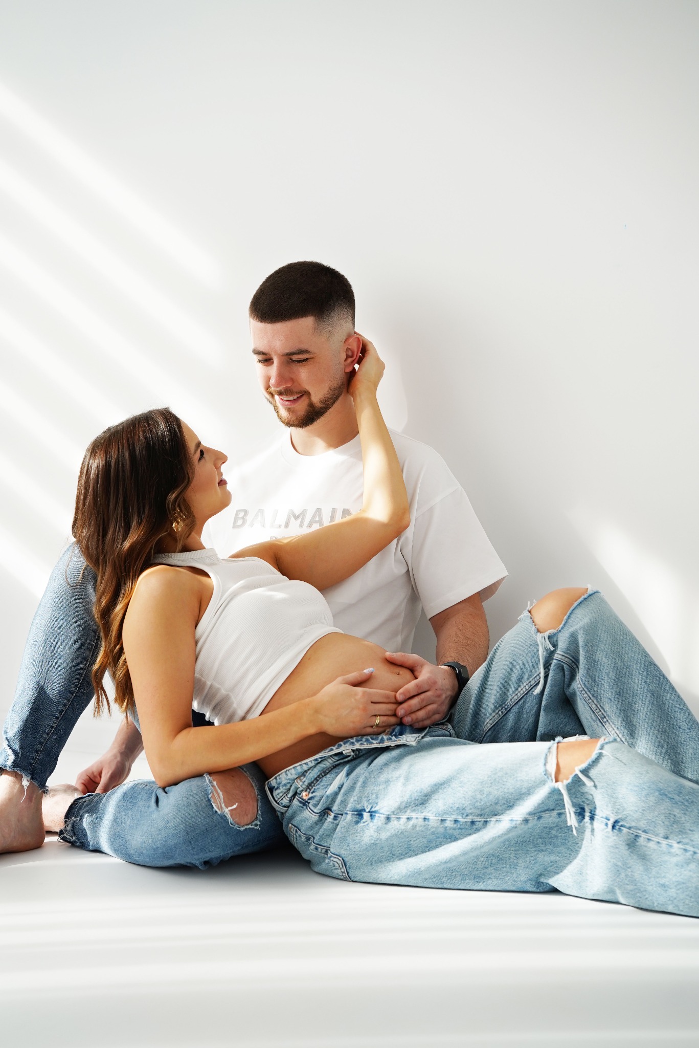 Uśmiechnięta para w jasnym studiu: przyszła mama w białym topie i jeansach z dziurami, czule dotyka twarzy partnera w białym t-shircie, dłonie na ciążowym brzuszku.