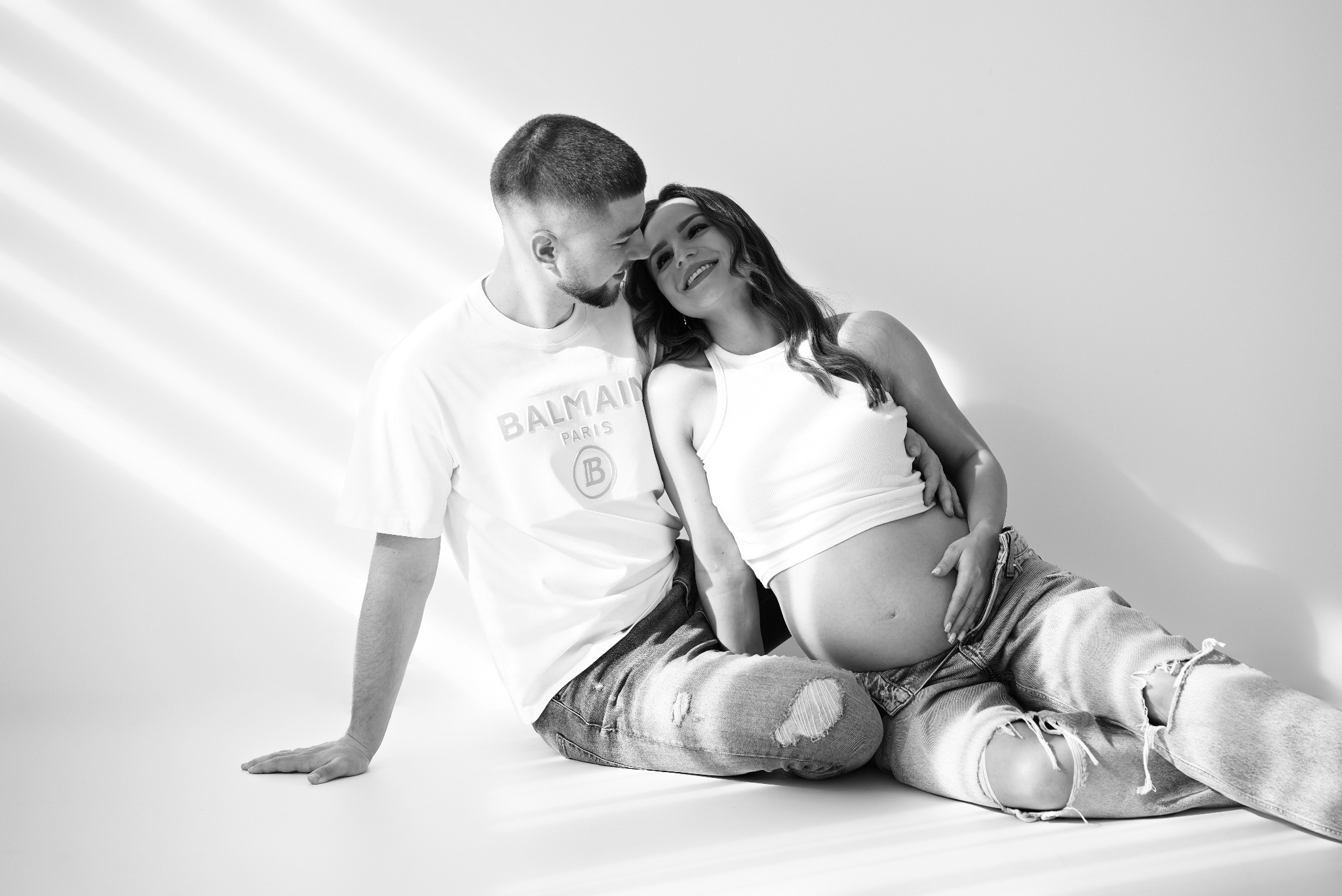 Czarno-białe zdjęcie pary: mężczyzna w koszulce Balmain obejmuje ciężarną kobietę w dżinsach z dziurami, siedzących na podłodze w jasnym świetle.