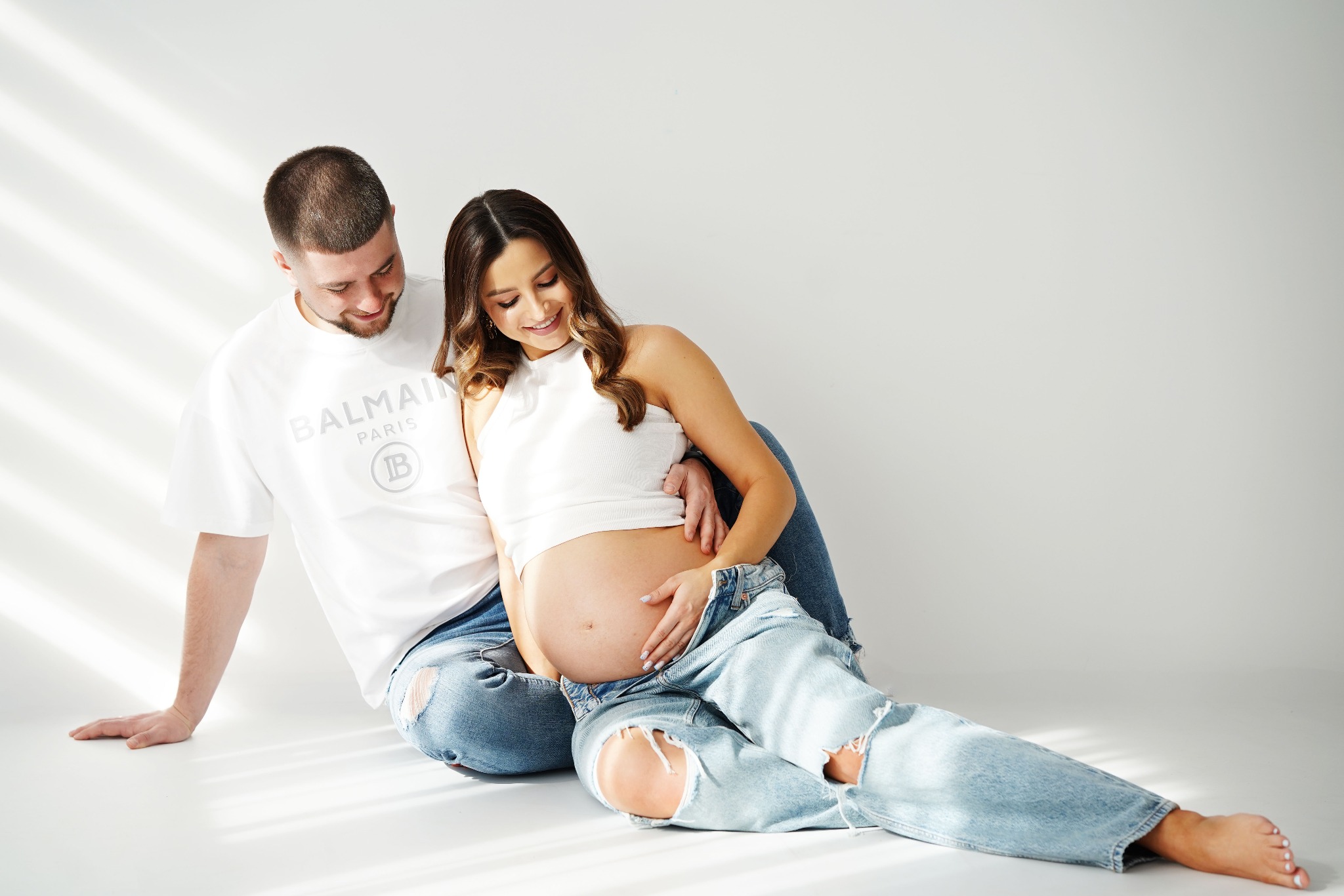 Uśmiechnięta para w jasnych jeansach siedzi na podłodze w studio. Kobieta w ciąży, mężczyzna obejmuje ją. Stonowana kolorystyka, miękkie światło.