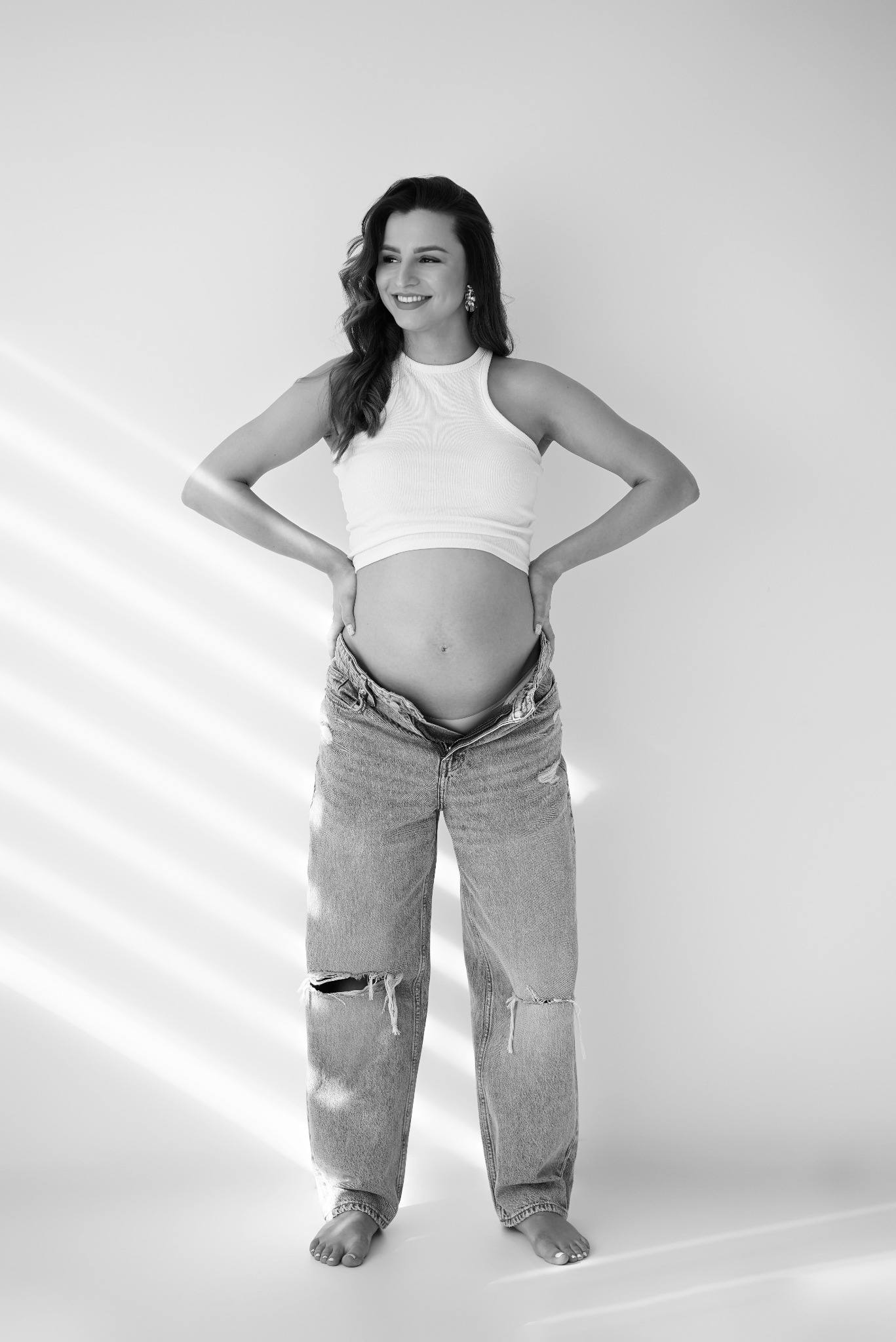 Czarno-białe zdjęcie uśmiechniętej kobiety w ciąży, w topie i rozpiętych jeansach, stojącej na tle jasnej ściany, z rękami na biodrach i delikatnym oświetleniem.