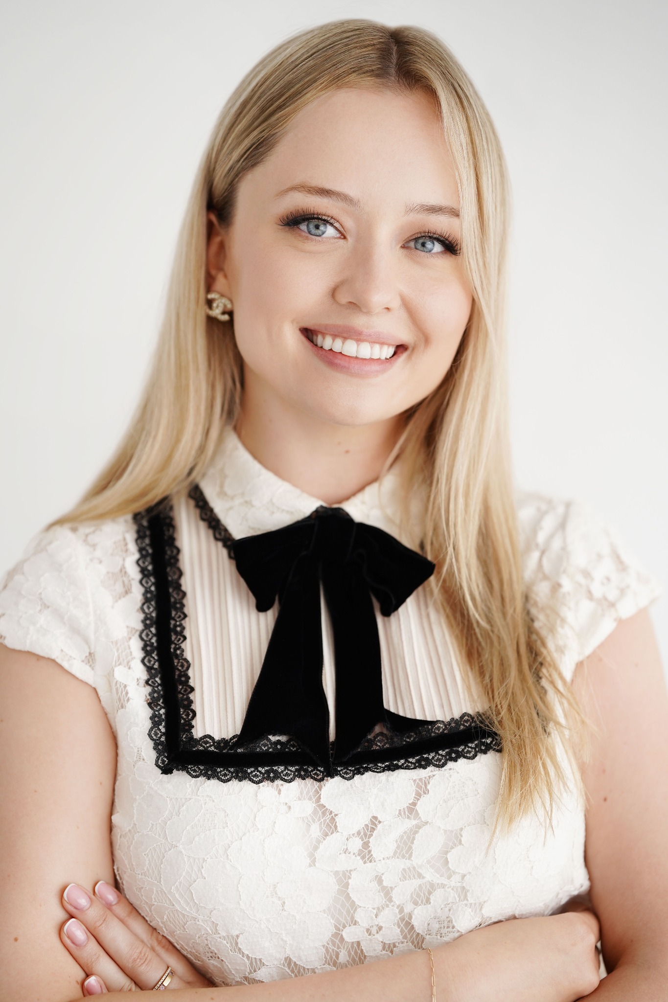 Uśmiechnięta blondynka w koronkowej bluzce z czarną kokardą i złotymi kolczykami Chanel na białym tle. Skrzyżowane ręce, delikatny makijaż.