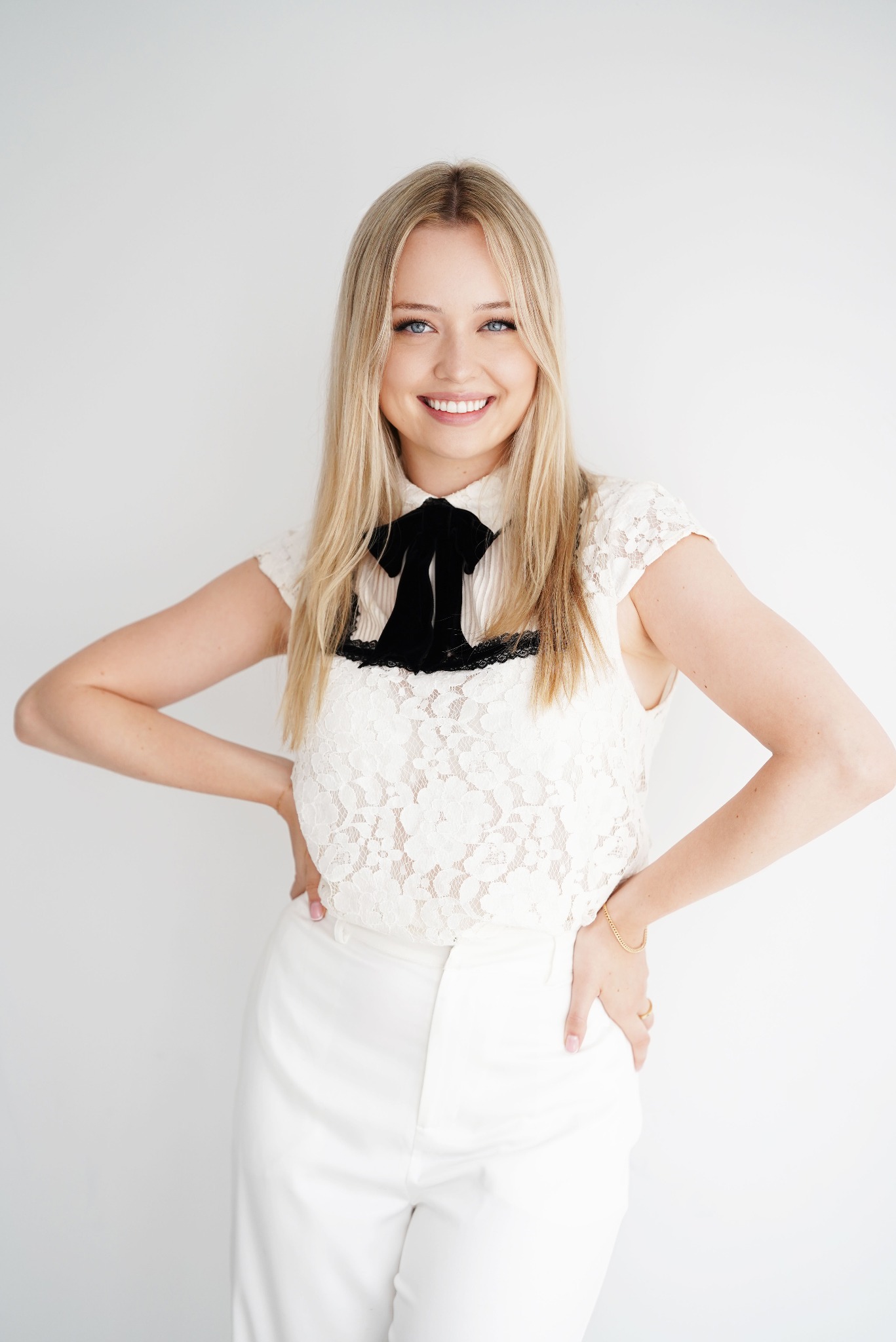 Uśmiechnięta blondynka w białym, koronkowym topie z czarną kokardą i białych spodniach, pozuje na jasnym tle, trzymając ręce na biodrach. Widoczny złoty łańcuszek na nadgarstku.
