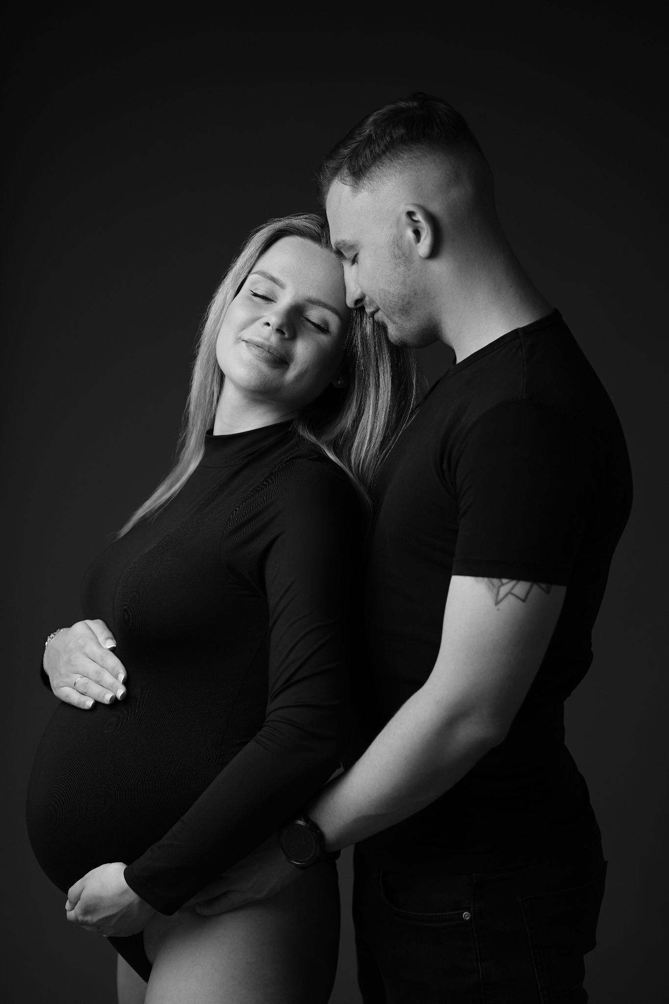 Czarno-białe zdjęcie kobiety w ciąży, obejmowanej przez mężczyznę. Ona w czarnym body, on w czarnym t-shircie. Delikatne światło podkreśla intymność i bliskość pary.