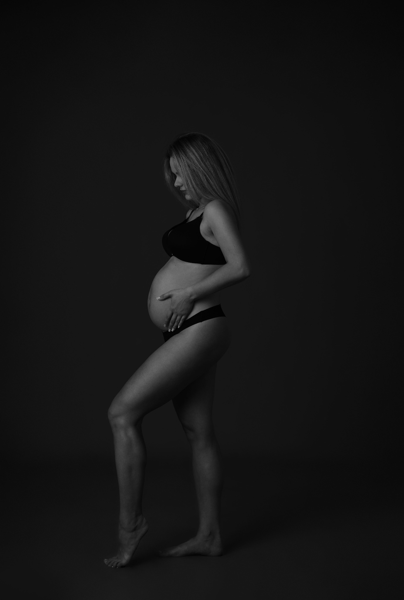 Monochromatyczne zdjęcie profilowe kobiety w ciąży, w bieliźnie, z rękami na brzuchu, na ciemnym tle. Subtelne oświetlenie podkreśla kształty.