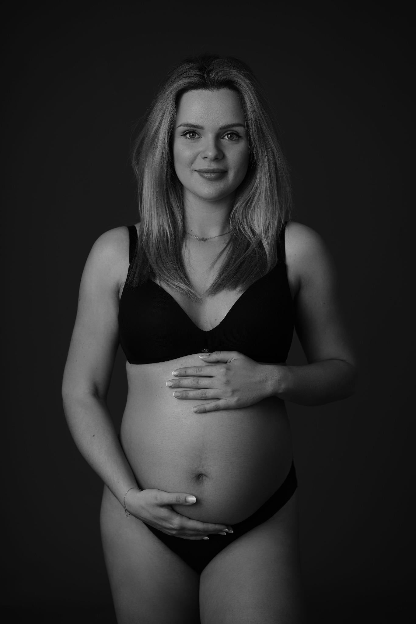 Czarno-białe zdjęcie portretowe kobiety w ciąży, w bieliźnie, z rękami na brzuchu, na ciemnym tle. Widoczny delikatny uśmiech i naturalne światło.