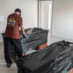 Pracownik firmy przeprowadzkowej w koszulce z logo firmy zabezpiecza meble folią stretch przed transportem w pustym pomieszczeniu.