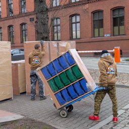 Piernik Przeprowadzki Toruń Transport Mebli Bagażówka - Dwóch pracowników w Toruniu rozładowuje meble po przeprowadzce. Jeden z nich transportuje regał na wózku, drugi zabezpiecza kartony. W tle budynek z czerwonej cegły.