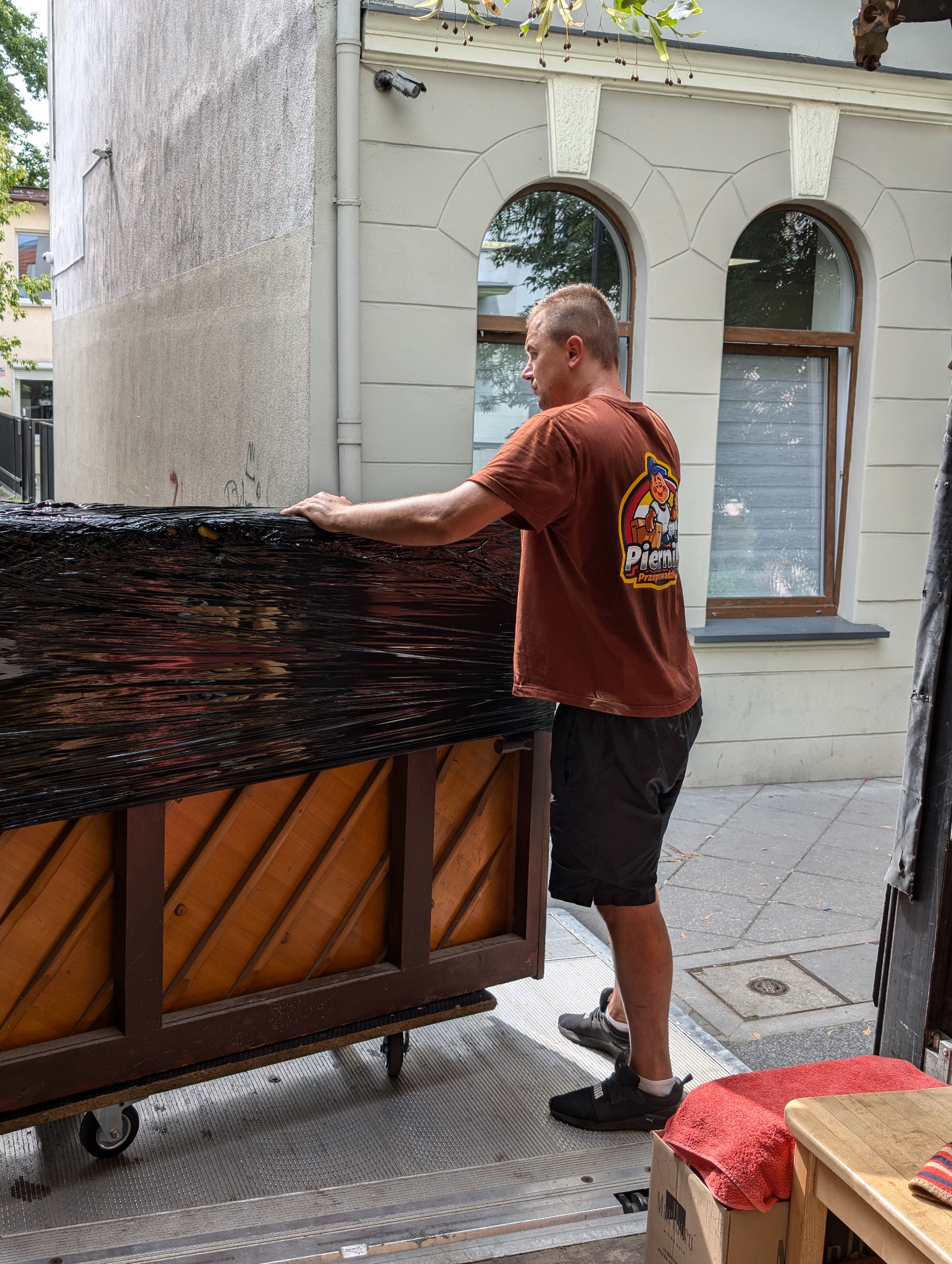 Mężczyzna w brązowej koszulce z logo firmy przeprowadzkowej zabezpiecza pianino folią stretch na platformie z kółkami przed budynkiem.