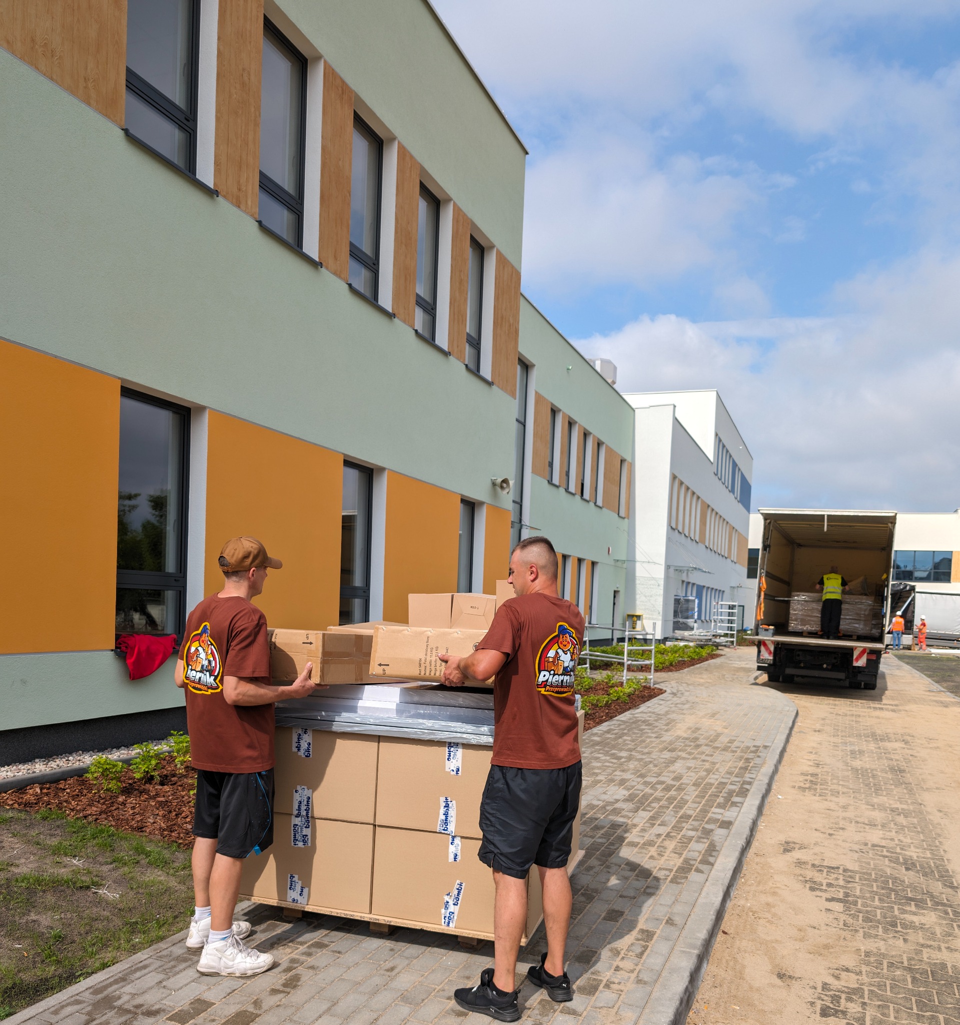 Dwóch mężczyzn w brązowych koszulkach z logo firmy transportowej przenosi kartony z palety przed nowoczesnym budynkiem w jasnych kolorach, w tle widoczna ciężarówka z otwartą paką i pracownikiem...