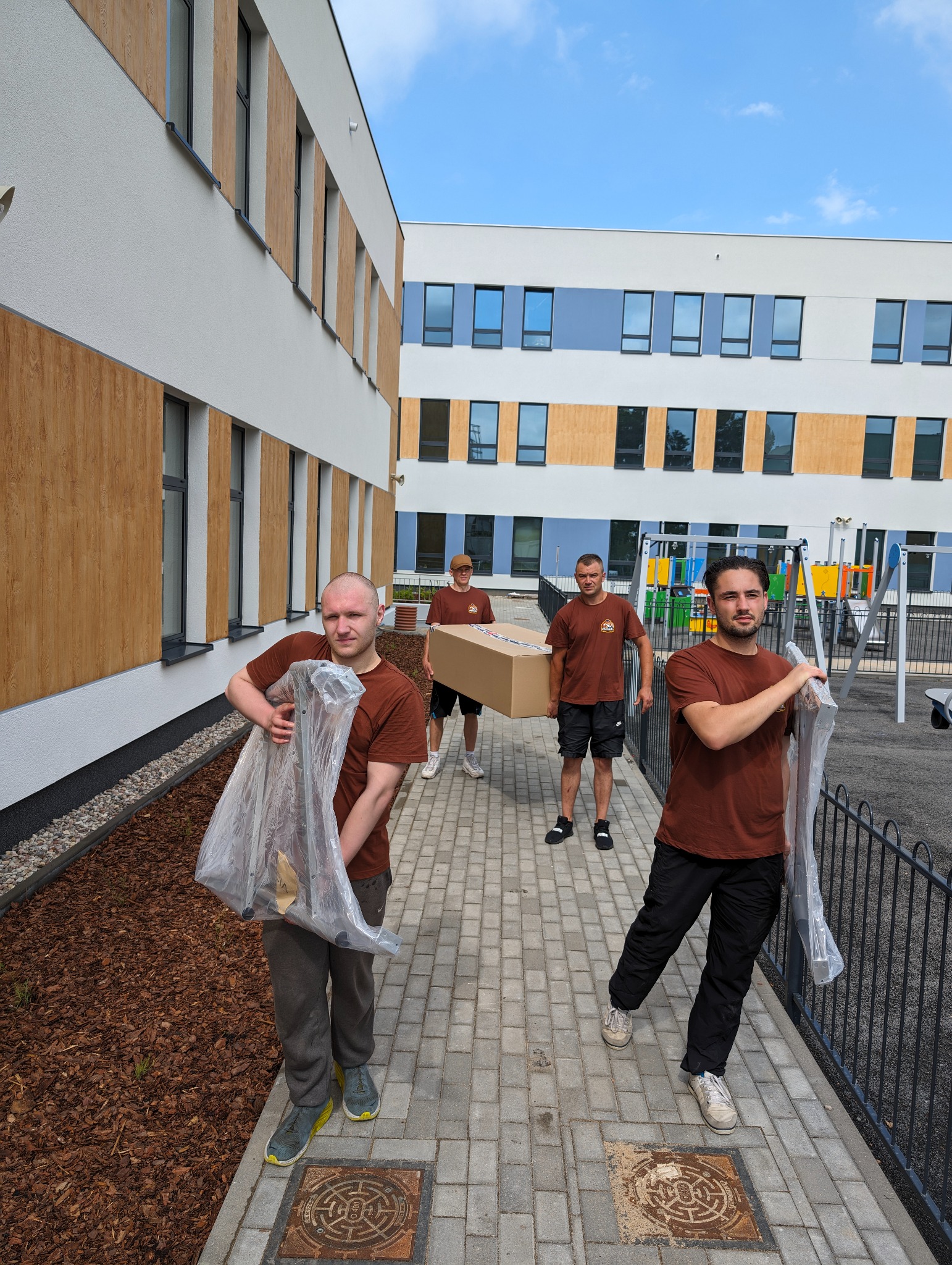 Czterech mężczyzn w brązowych koszulkach przenosi kartonowe pudło i meble zapakowane w folię na tle nowego budynku z placem zabaw.