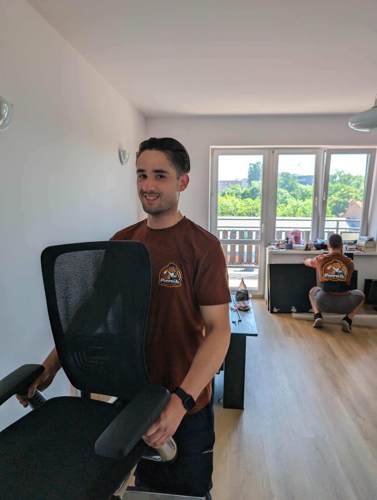 Uśmiechnięty mężczyzna w brązowej koszulce z logo firmy przeprowadzkowej, niosący czarne krzesło biurowe na tle jasnego mieszkania podczas przeprowadzki, drugi pracownik w tle.