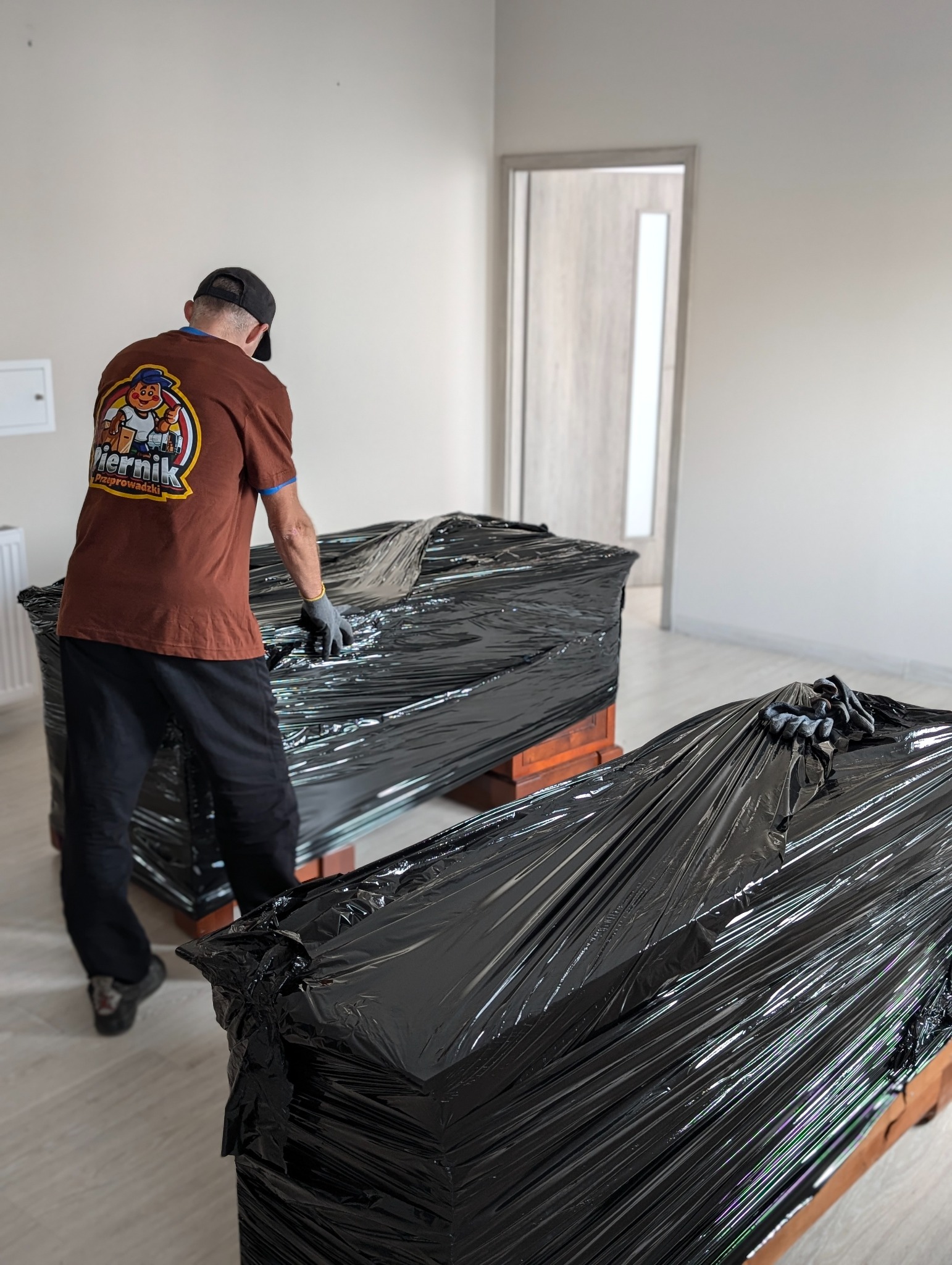 Pracownik firmy przeprowadzkowej w koszulce z logo firmy zabezpiecza meble folią stretch przed transportem w pustym pomieszczeniu.