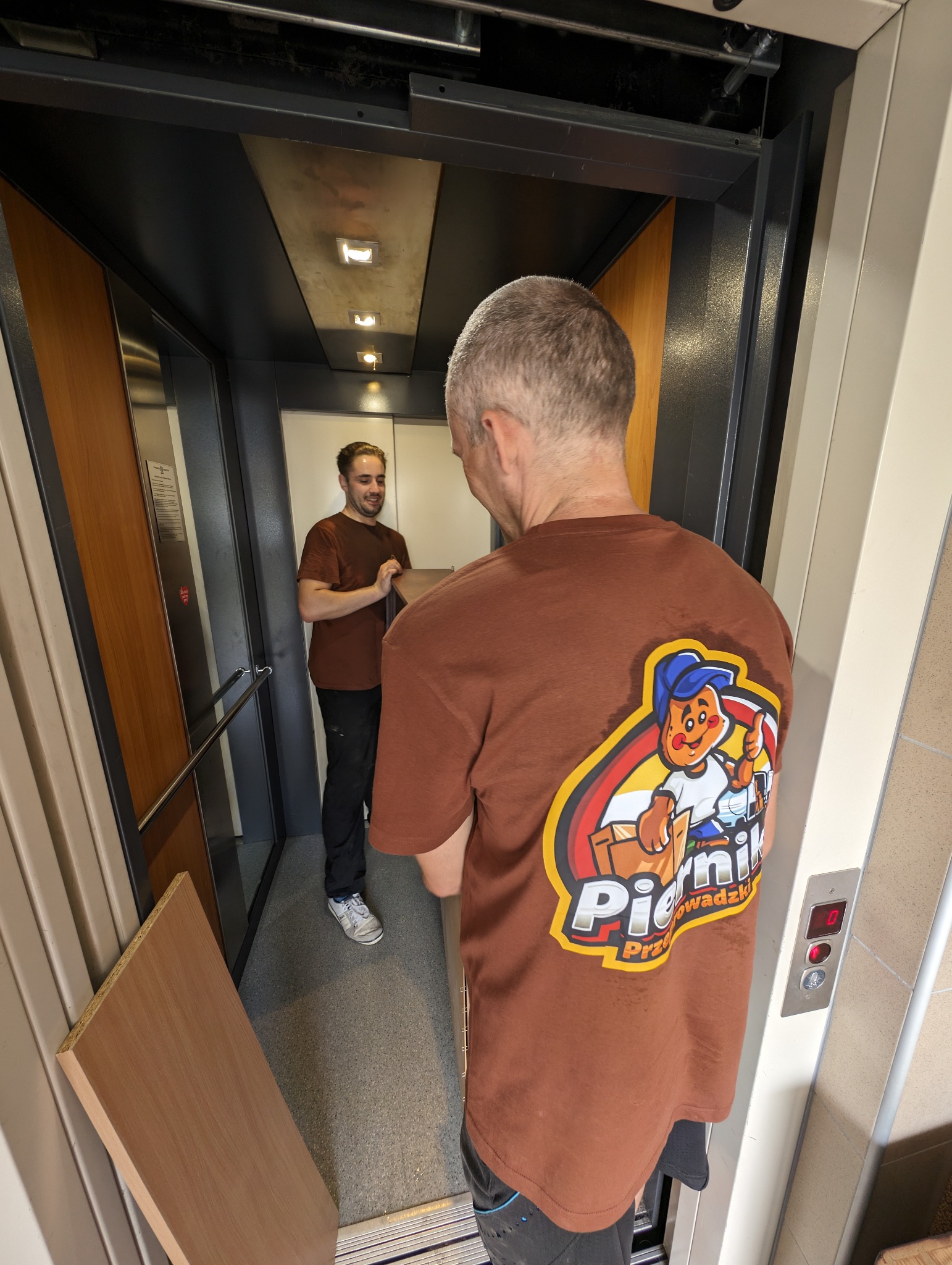 Dwóch mężczyzn w brązowych koszulkach z logo firmy transportowej w windzie, przenoszą płytę meblową.