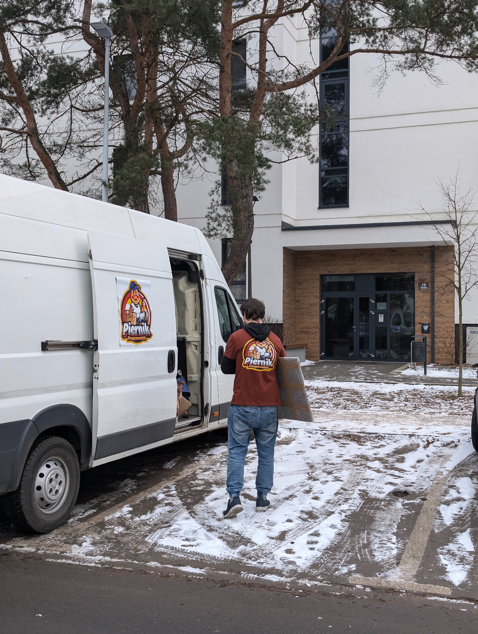 Pracownik firmy przeprowadzkowej 'Piernik' wnosi zabezpieczony dywan do busa na tle nowoczesnego budynku w zimowej scenerii.