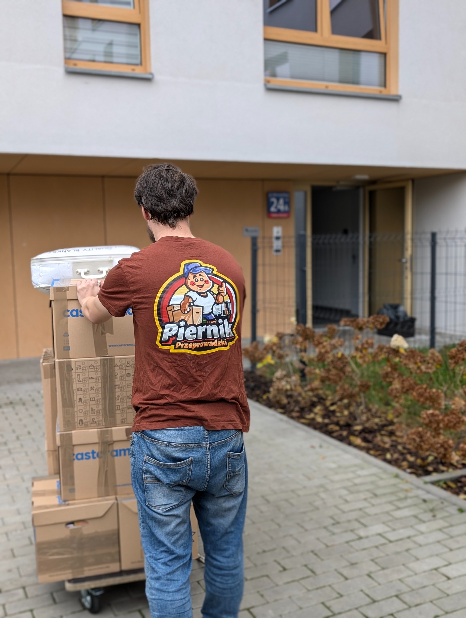 Mężczyzna w brązowej koszulce z logo firmy przeprowadzkowej 'Piernik', transportujący kartony na wózku przed budynkiem.