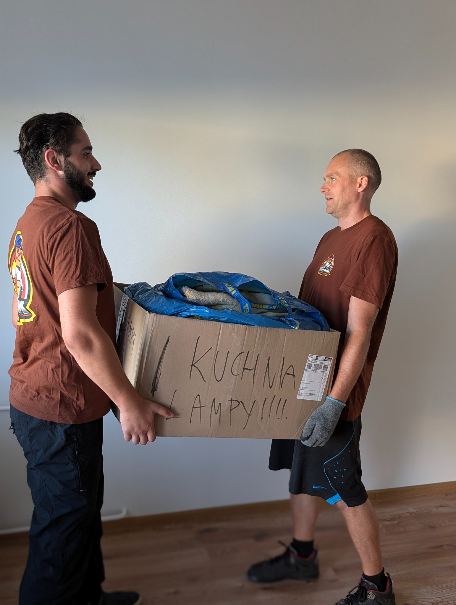 Dwóch mężczyzn w brązowych koszulkach z logo firmy transportowej niesie karton z napisem 'KUCHNIA LAMPY!!!!' wypełniony przedmiotami w niebieskiej torbie IKEA.