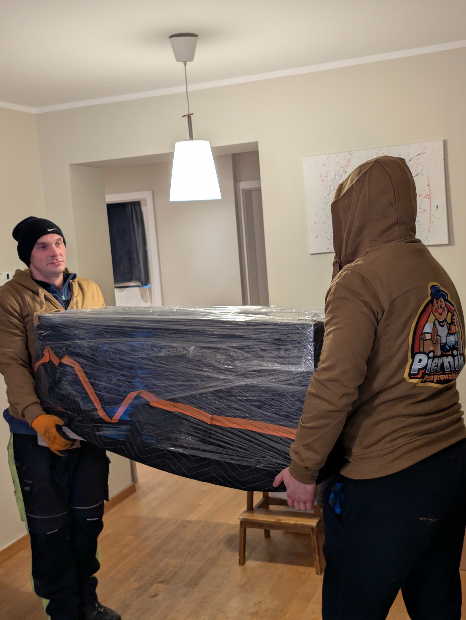 Dwóch mężczyzn w trakcie przeprowadzki niesie zapakowany mebel w folii stretch i kocu transportowym. Widoczny fragment wnętrza mieszkania z drewnianą podłogą.