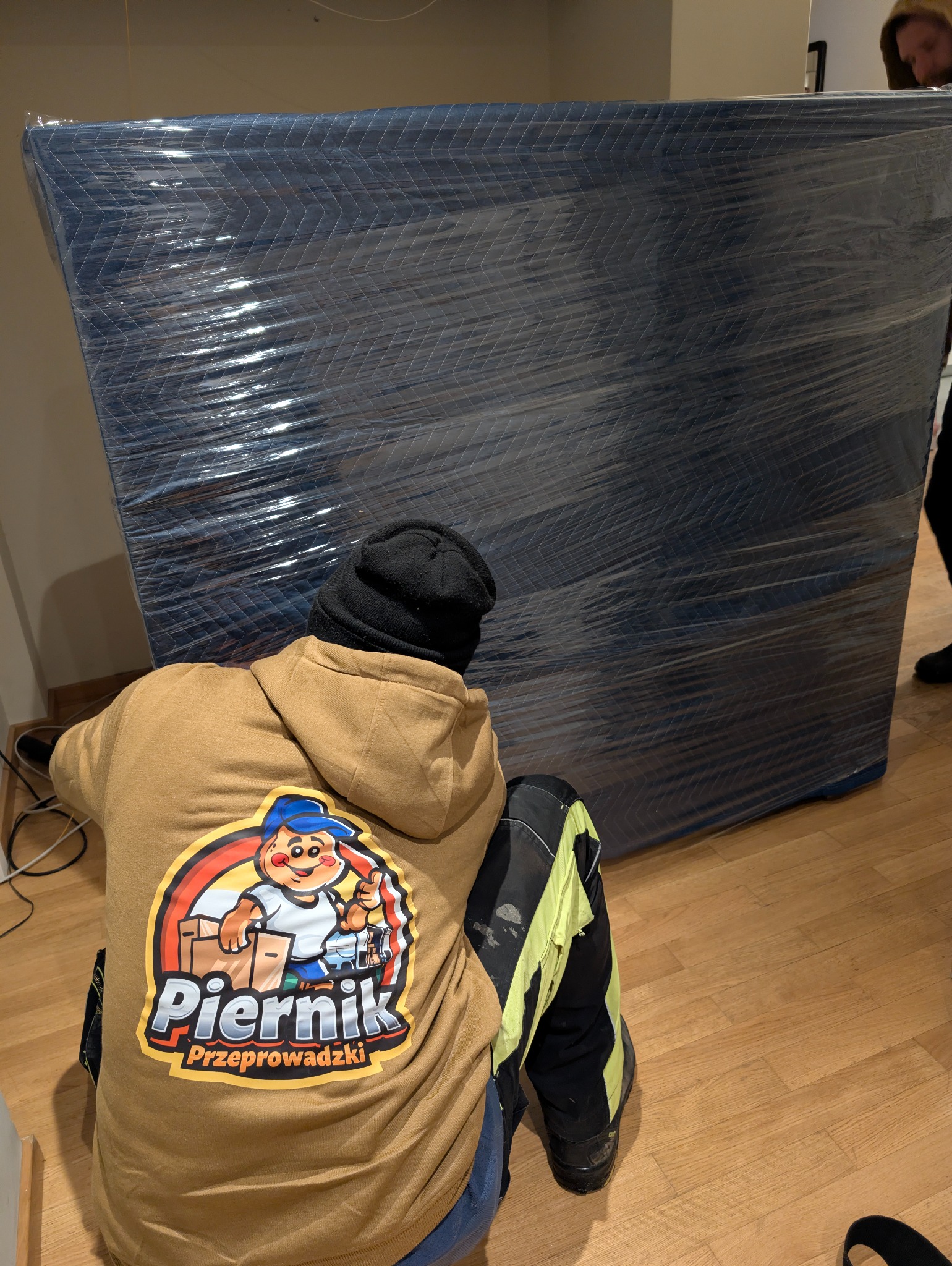 Pracownik firmy przeprowadzkowej w firmowej bluzie klęczy obok mebla zabezpieczonego folią i kocem transportowym. Widoczny fragment podłogi i ściany.