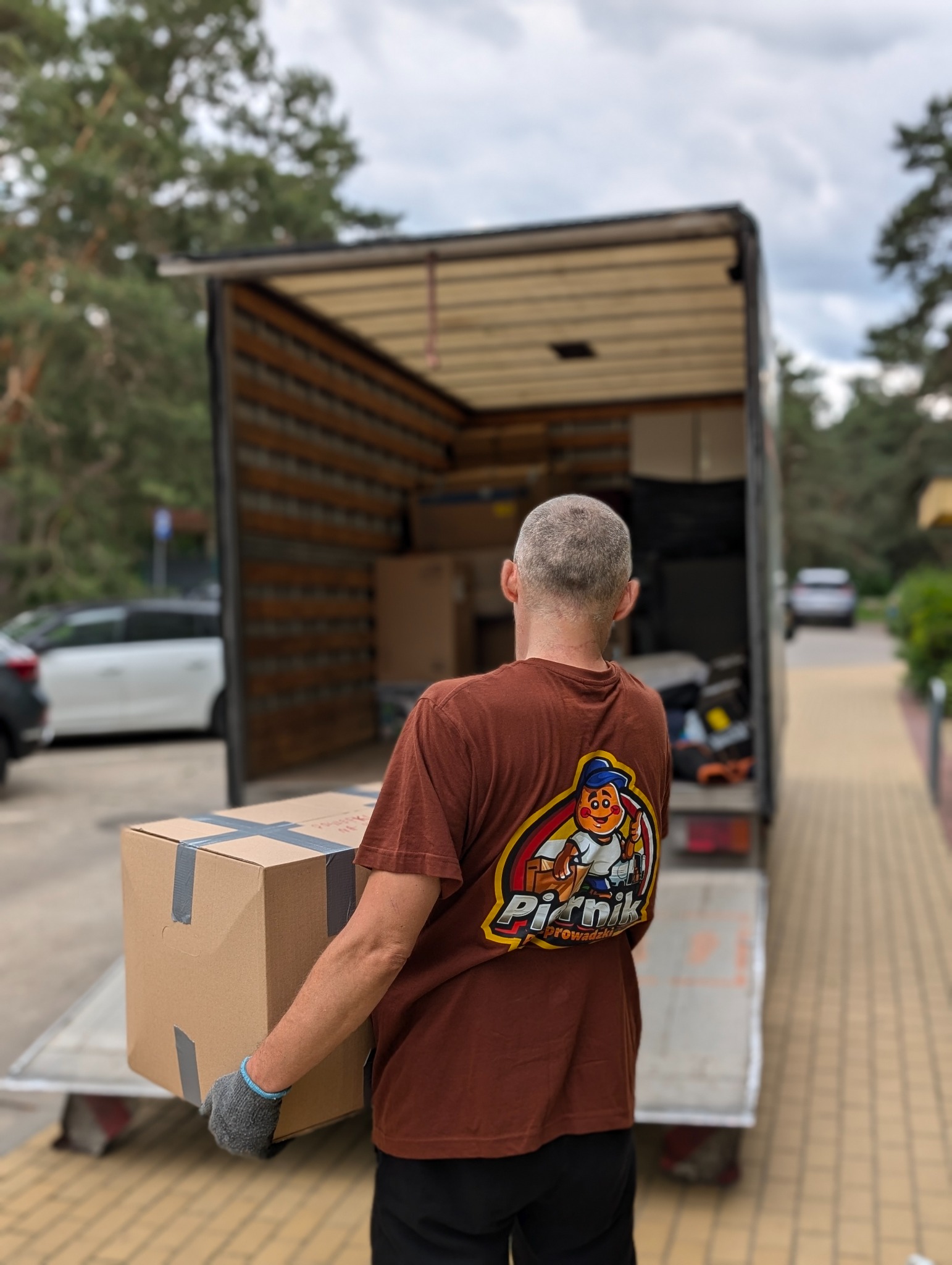 Pracownik firmy przeprowadzkowej z kartonem na tle otwartej ciężarówki. Na koszulce logo firmy 'Pizdnik Przeprowadzki'.