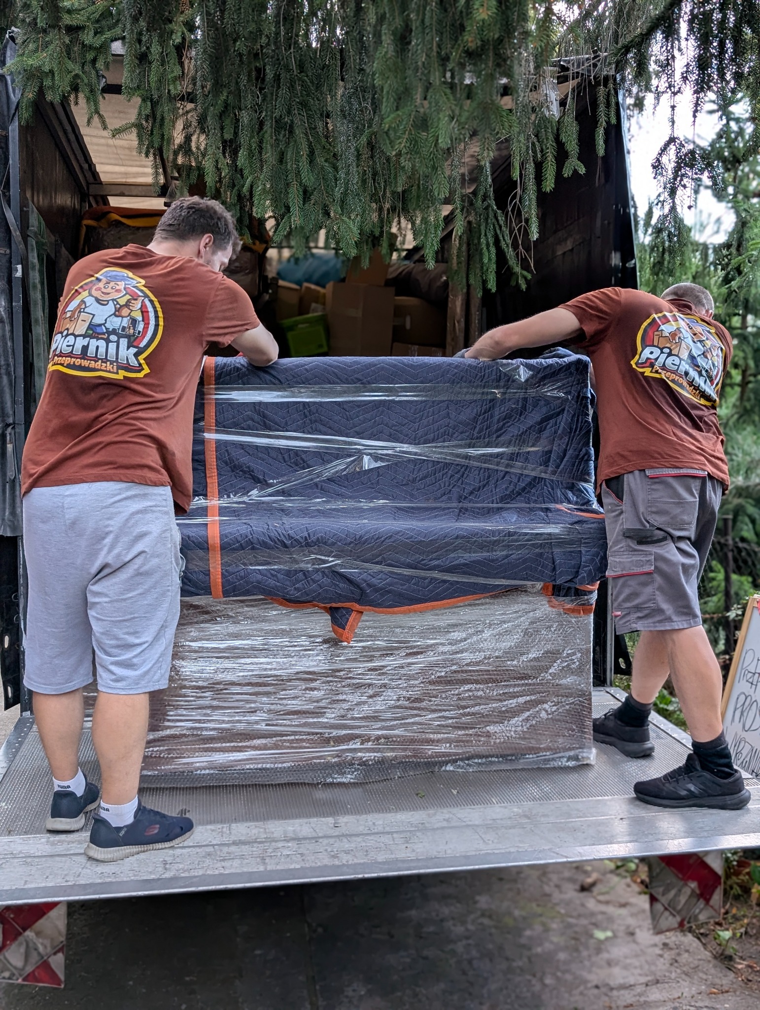 Dwóch mężczyzn w koszulkach z logo firmy przeprowadzkowej umieszcza zabezpieczony mebel w samochodzie dostawczym. Widoczne drzewa nad pojazdem.