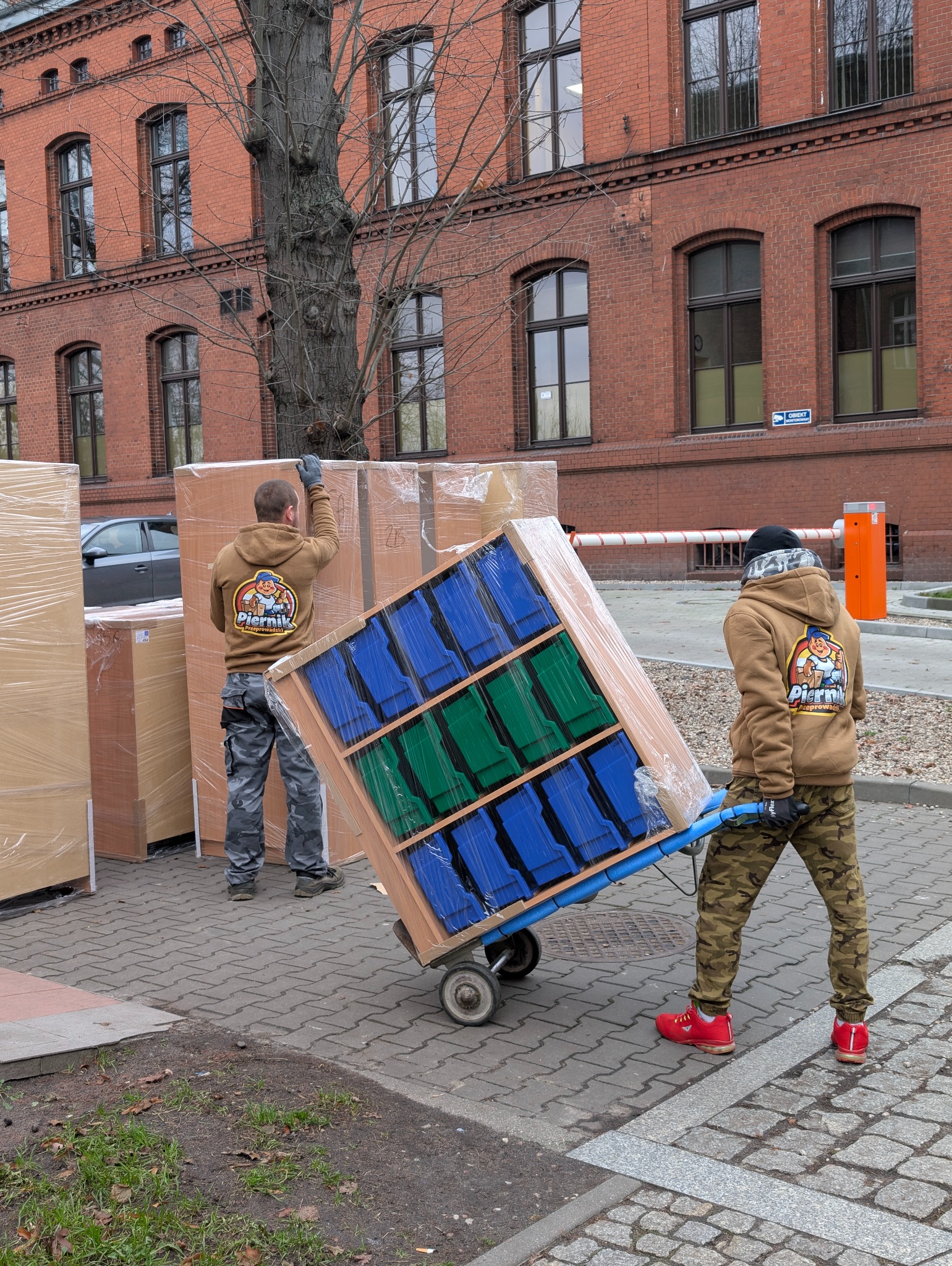 Dwóch pracowników w Toruniu rozładowuje meble po przeprowadzce. Jeden z nich transportuje regał na wózku, drugi zabezpiecza kartony. W tle budynek z czerwonej cegły.