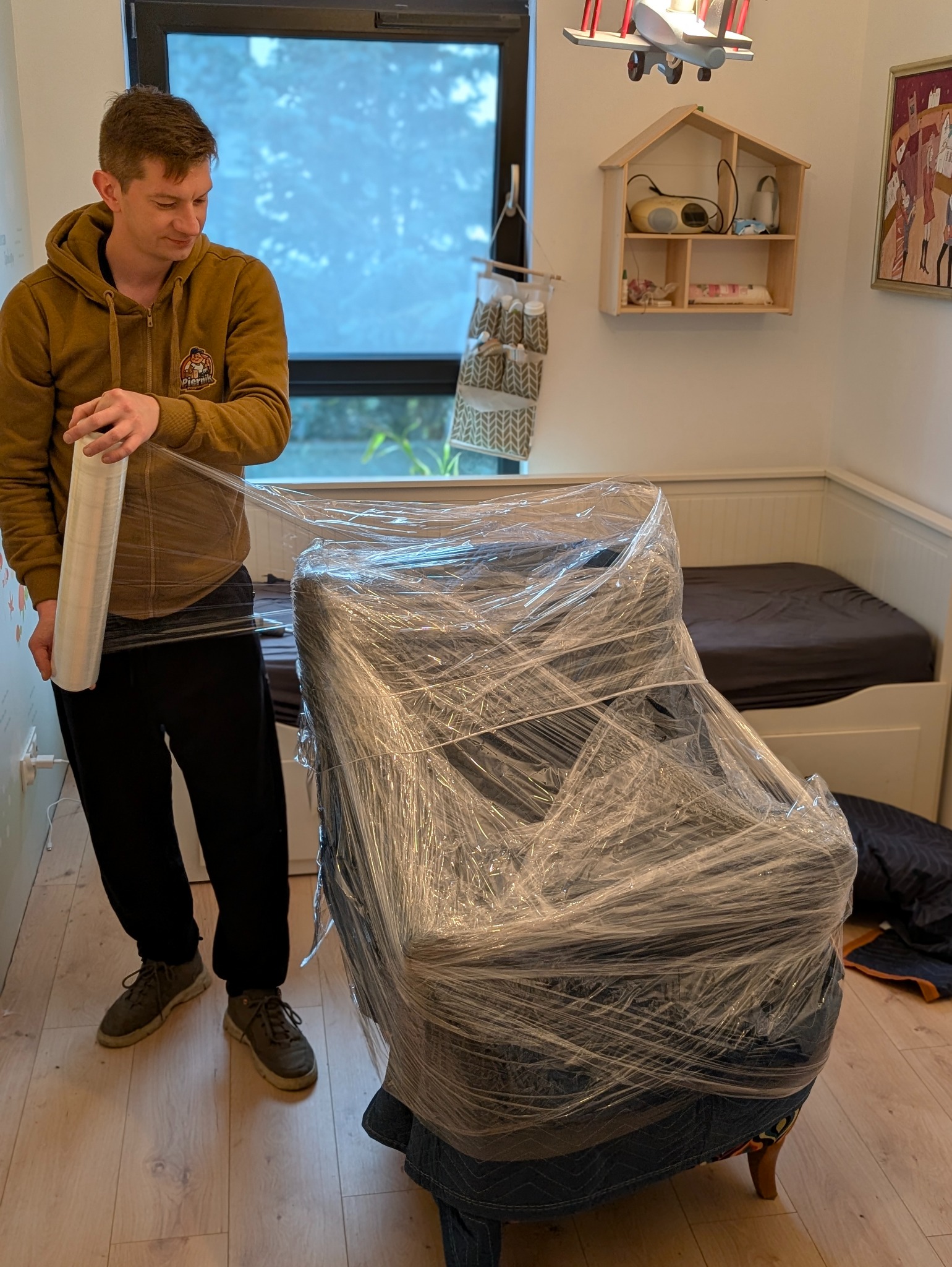 Mężczyzna owija fotel folią stretch w pokoju z łóżkiem i dekoracjami. Przygotowanie mebla do przeprowadzki, zabezpieczenie przed uszkodzeniem.