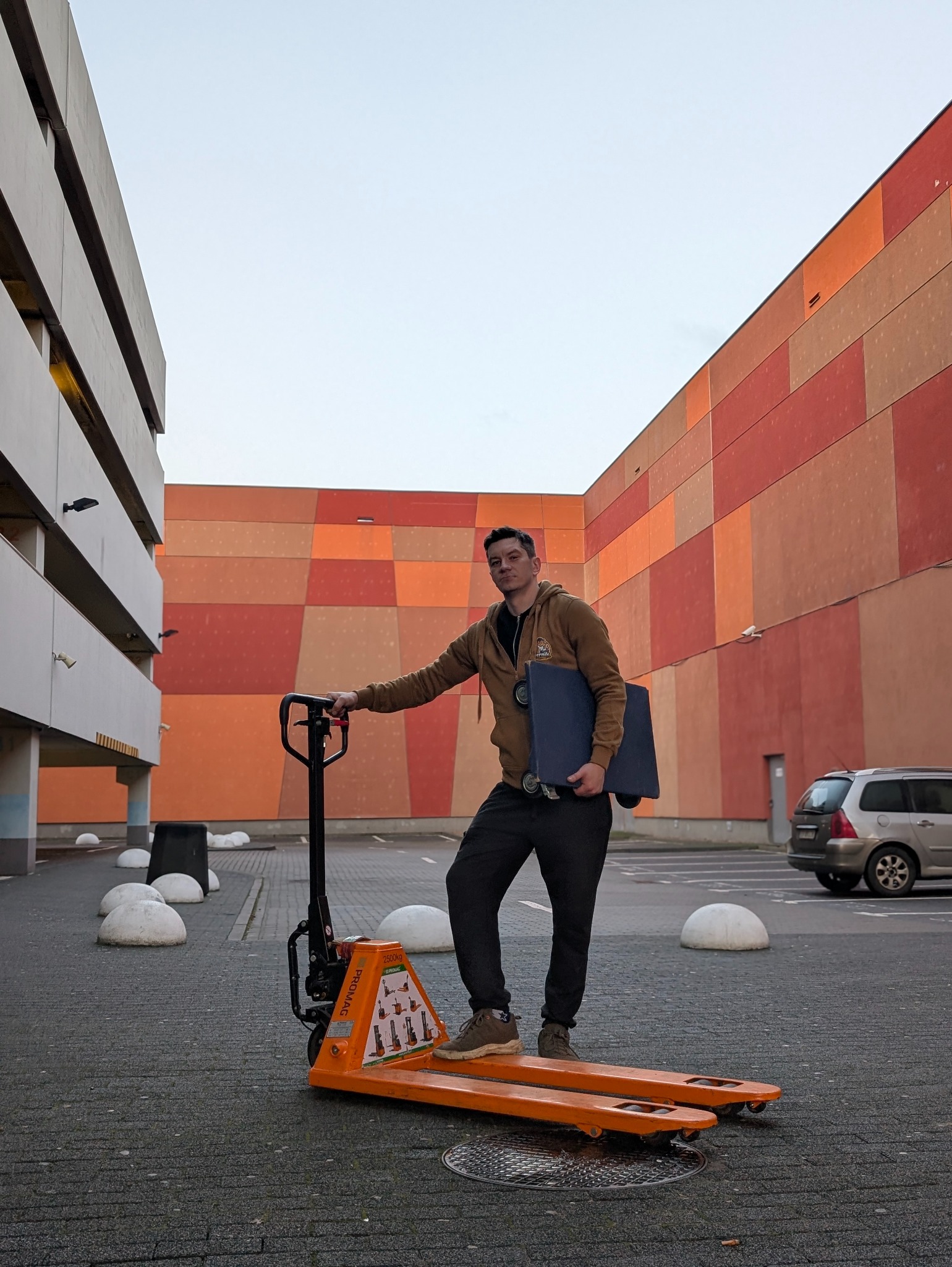 Mężczyzna z wózkiem paletowym na tle parkingu wielopoziomowego. Wózek pomarańczowy, budynek w odcieniach pomarańczy i szarości. Ujęcie z dołu, pochmurne niebo.
