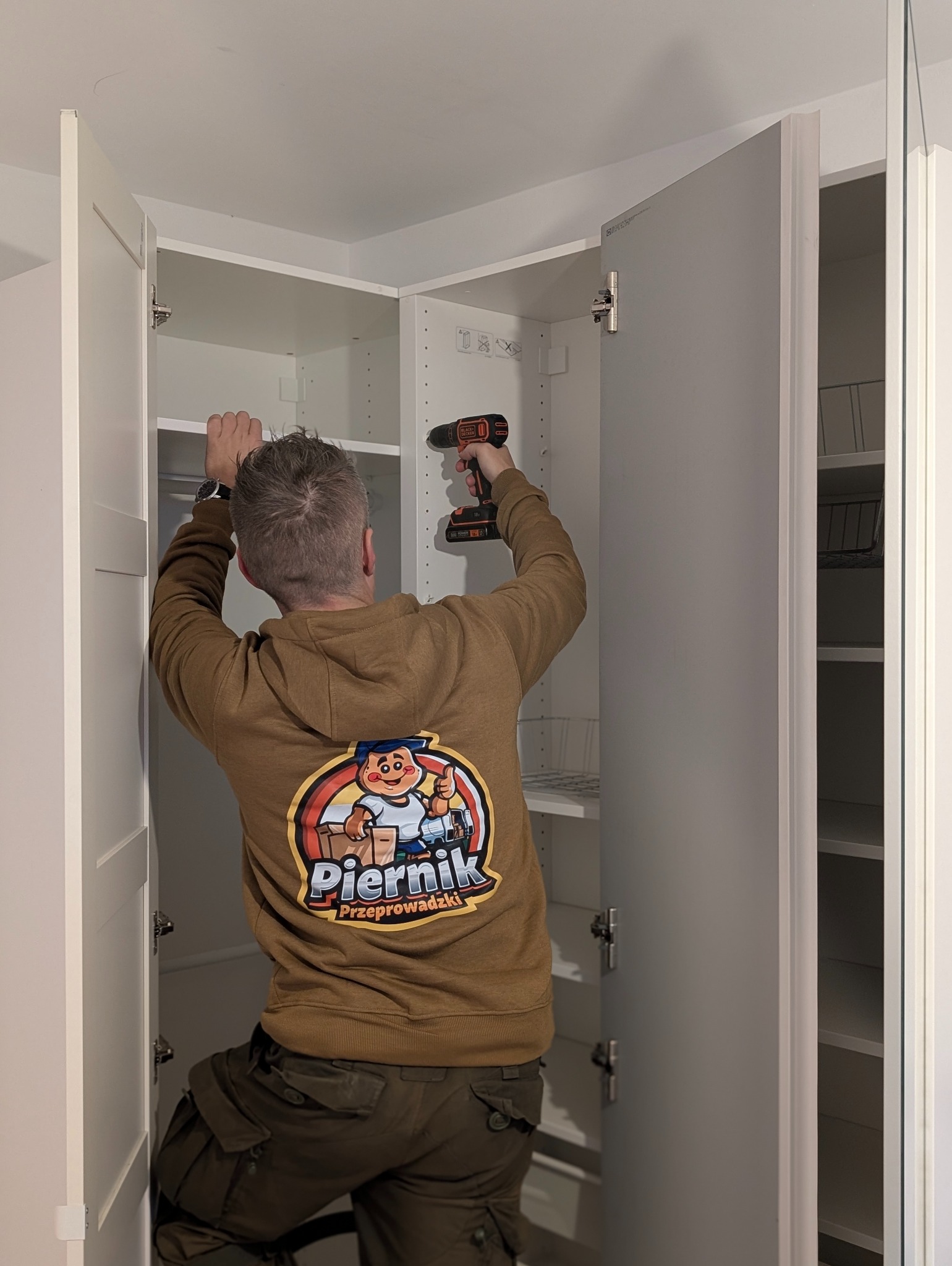 Pracownik firmy przeprowadzkowej montuje szafę wnękową w jasnym pomieszczeniu. Widoczny tył osoby w bluzie z logo firmy Piernik Przeprowadzki i wkrętarka.
