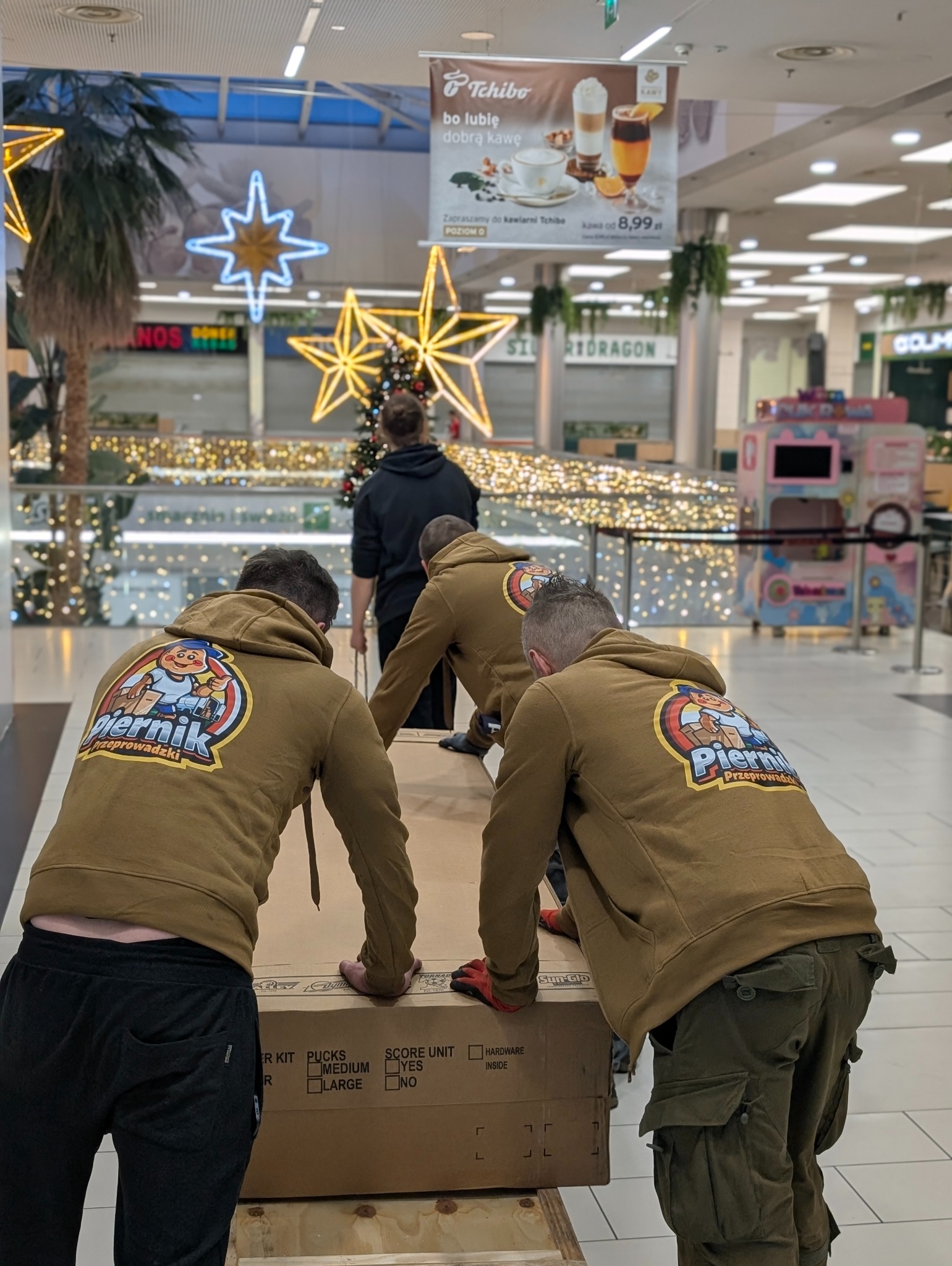 Ekipa przeprowadzkowa 'Piernik' w brązowych bluzach z logo firmy transportuje duży karton w centrum handlowym, w tle dekoracje świąteczne i reklama kawiarni.