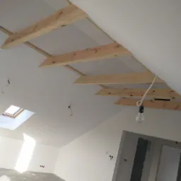 Wnętrze poddasza w Pniewach w trakcie prac wykończeniowych: widoczne drewniane elementy konstrukcyjne sufitu, okno dachowe wpuszczające jasne światło oraz surowe ściany przygotowane do dalszych prac.