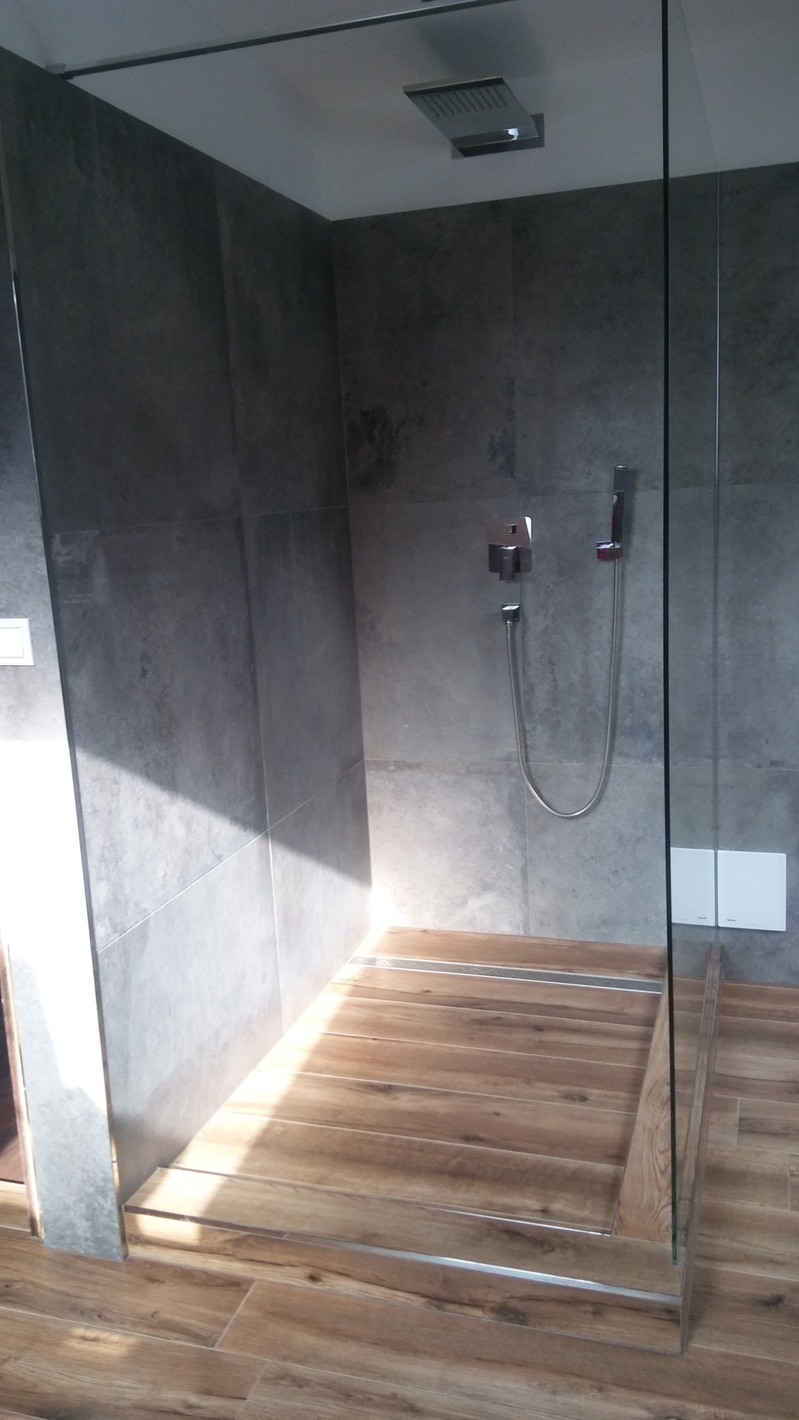 Nowoczesna kabina prysznicowa z szarymi płytkami na ścianach i drewnopodobną podłogą z odpływem liniowym.
