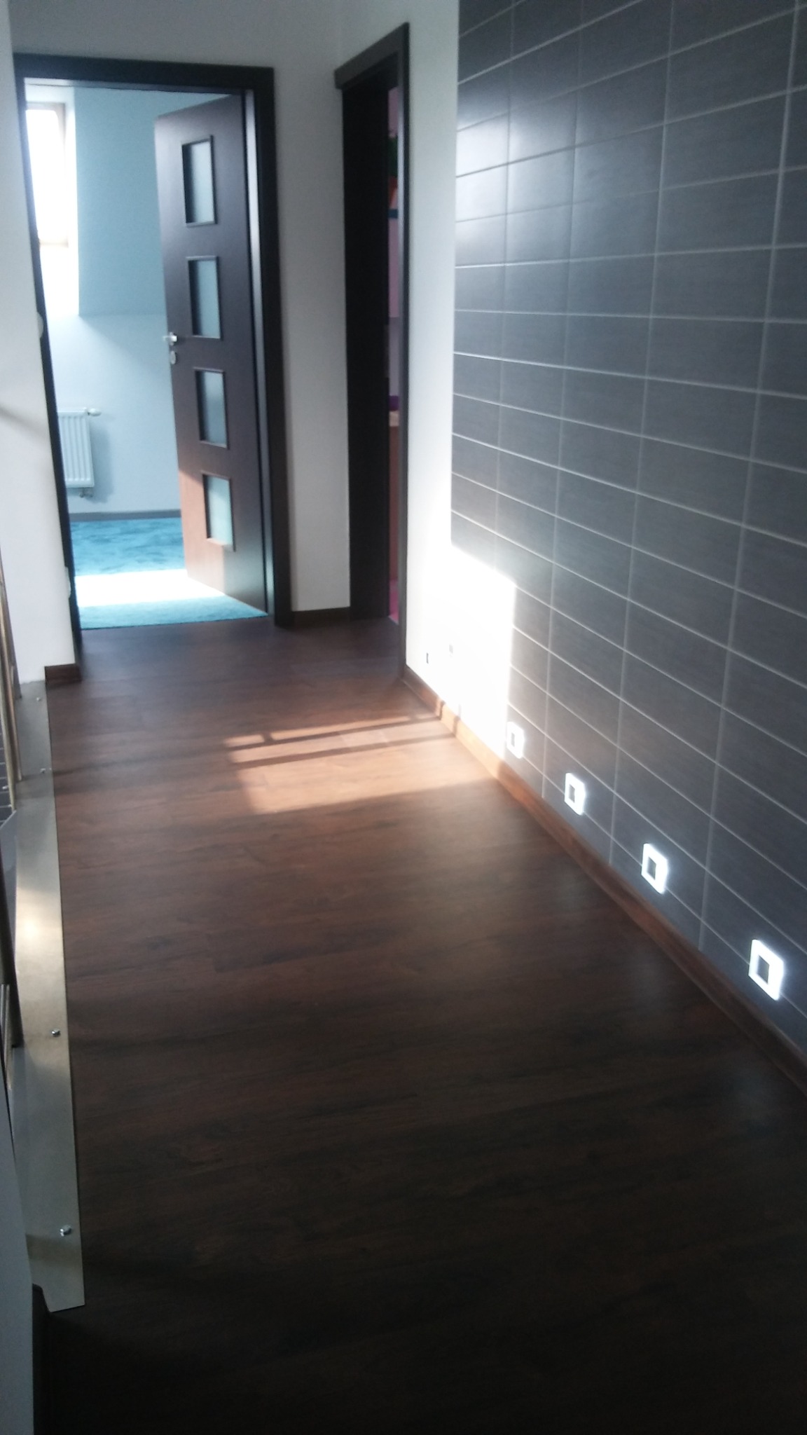Korytarz z ciemną drewnopodobną podłogą, szarą ścianą wyłożoną płytkami i punktowym oświetleniem podłogowym. W tle widoczne otwarte drzwi i jasne pomieszczenie.