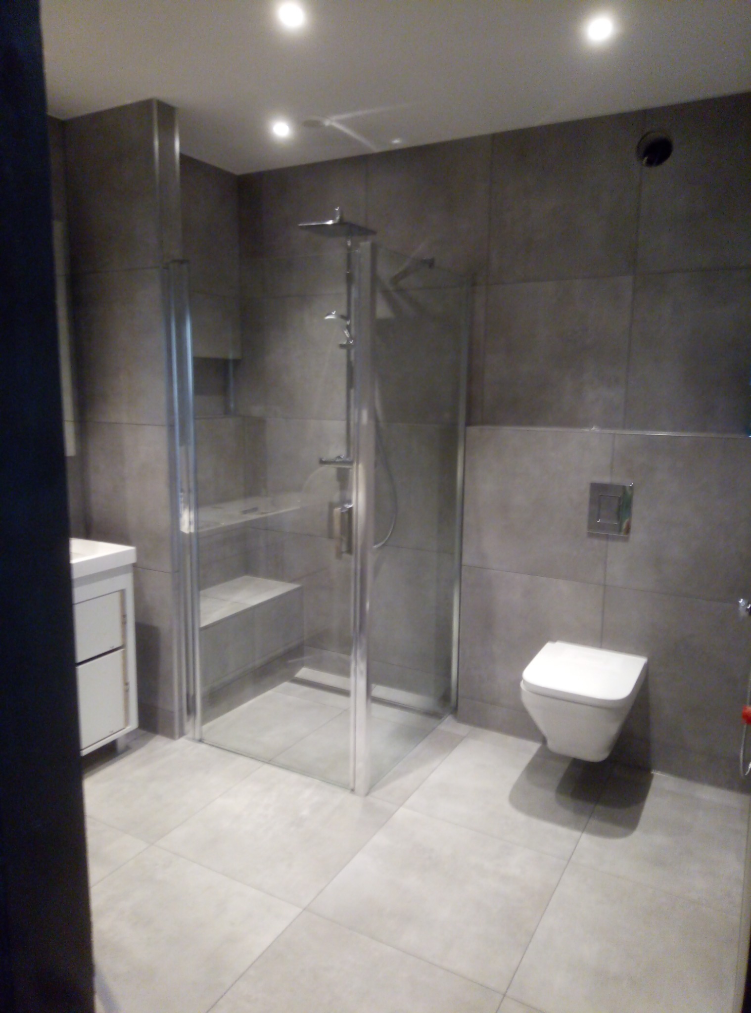 Nowoczesna łazienka z szarymi płytkami, kabiną prysznicową typu walk-in z półkami, podwieszaną toaletą i białą szafką pod umywalką.