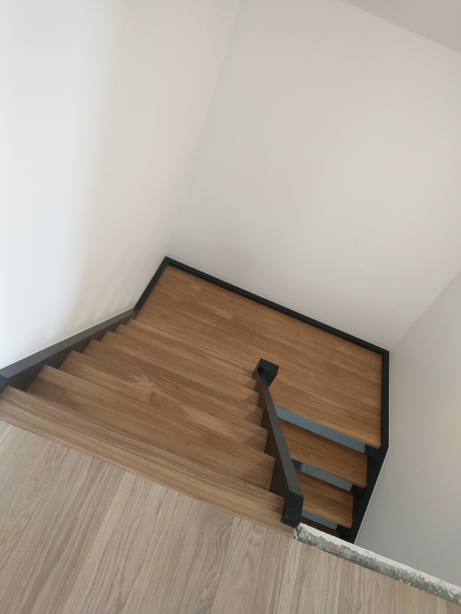 Schody drewniane z czarnymi metalowymi elementami, widok z góry, oświetlone naturalnym światłem, z widocznymi śladami prac wykończeniowych na podłodze.