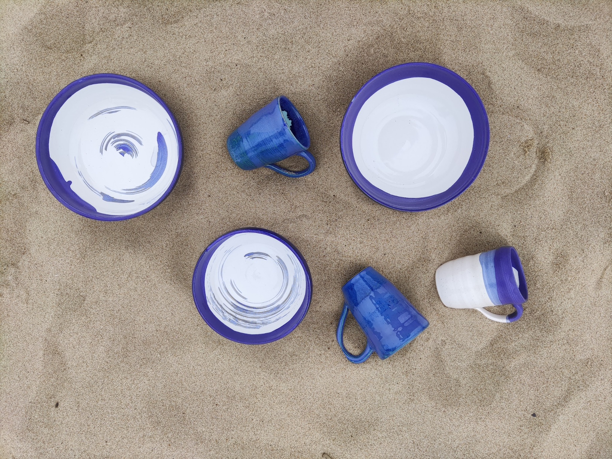 Ręcznie robione ceramiczne talerze i kubki z niebieskimi akcentami, ułożone na piasku. Widok z góry.