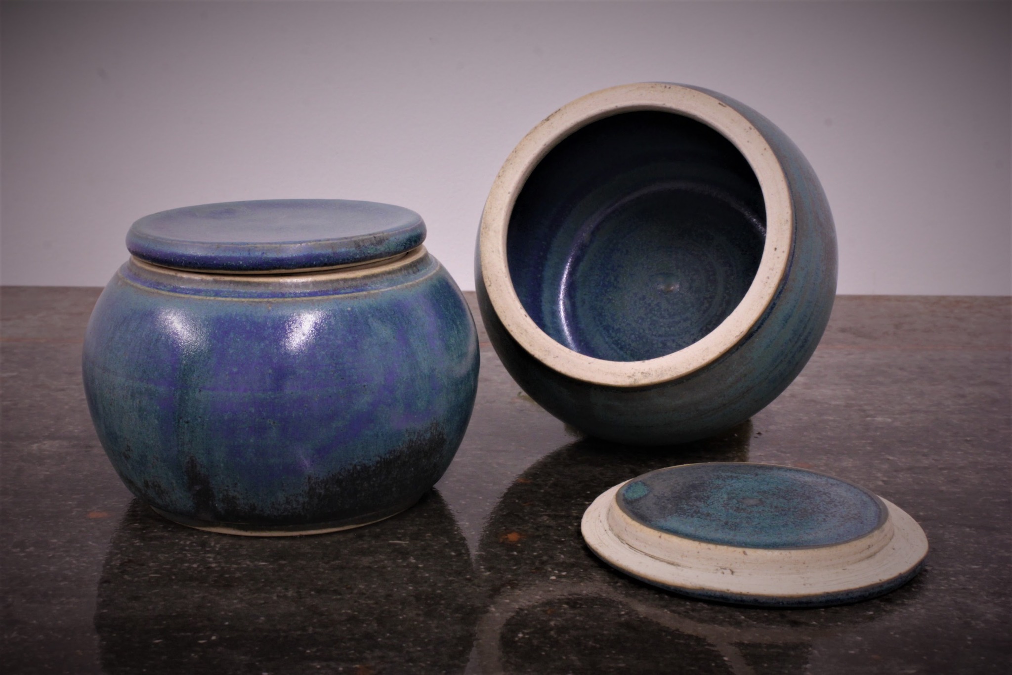 Dwa ceramiczne pojemniki z pokrywkami w odcieniach niebieskiego i turkusu, jeden zamknięty, drugi otwarty, leżąca obok pokrywka, na ciemnym blacie.