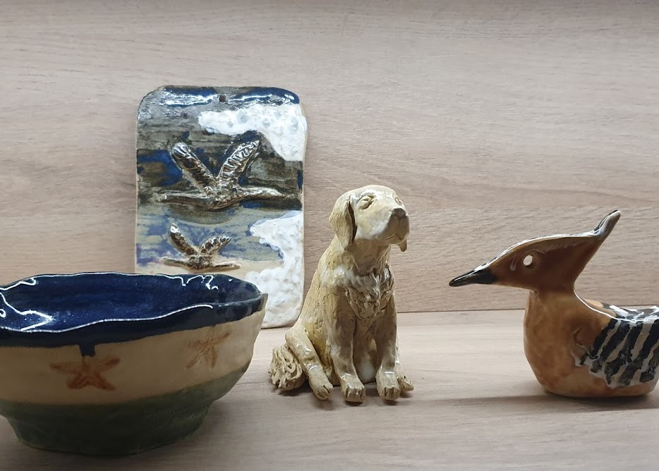 Ręcznie wykonane ceramiczne figurki: miseczka z motywem rozgwiazd, płytka z motywem ptaków w locie, pies i ptak dudek, ustawione na drewnianej półce.