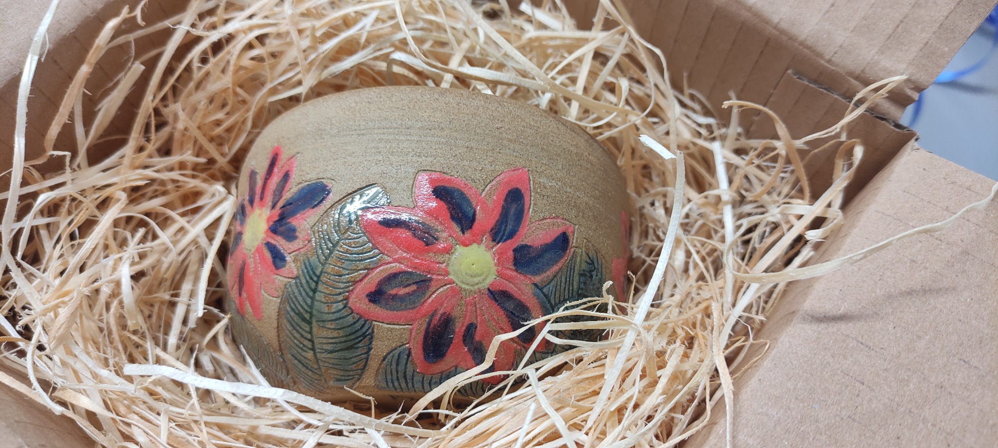 Ceramiczna doniczka z ręcznie malowanymi czerwonymi kwiatami i zielonymi liśćmi, umieszczona w kartonowym pudełku wypełnionym siankiem.