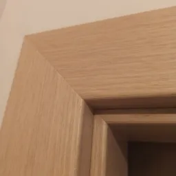 Fragment drewnianej ościeżnicy drzwiowej, ukazujący połączenie górnej i bocznej części ramy z widoczną strukturą drewna i delikatnym zaokrągleniem krawędzi.