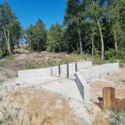 Wylany fundament pod budynek z widocznymi stalowymi elementami szalunku w gruncie, otoczony drzewami i ziemią.