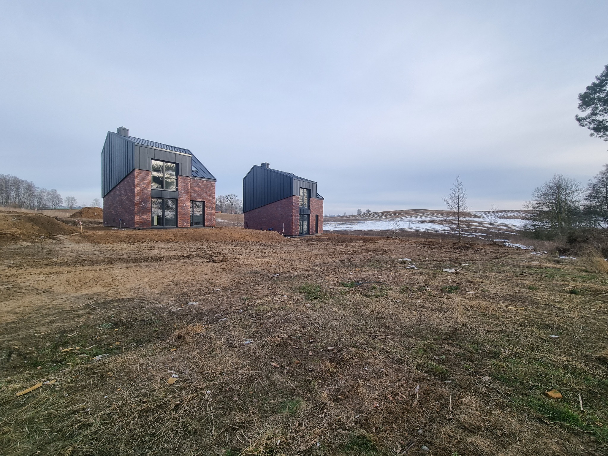 Dwa nowoczesne domy z cegły klinkierowej z czarnym dachem na nierównym terenie z resztkami śniegu w tle.