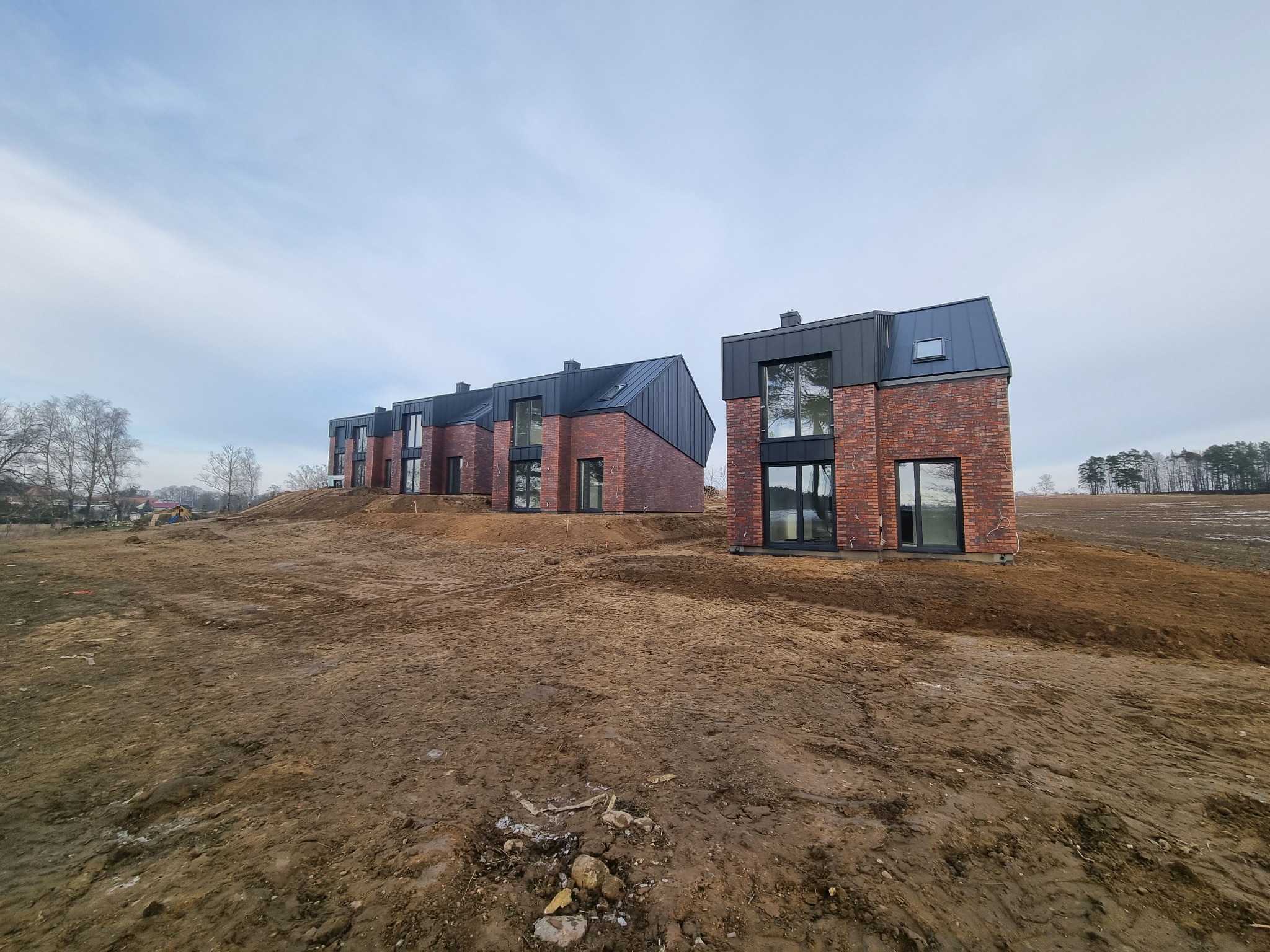 Trzy nowoczesne domy z czerwonej cegły z czarnymi dachami, jeszcze w trakcie budowy, na nierównym, przygotowanym terenie.